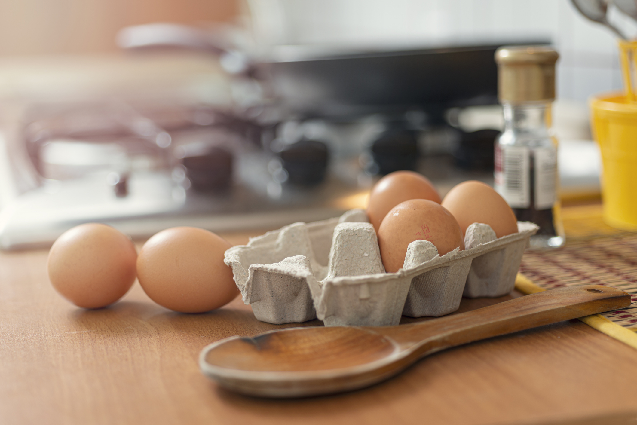 Eggs in a carton and a wooden spoon on a kitchen countertop, with a stove and spice container in the background