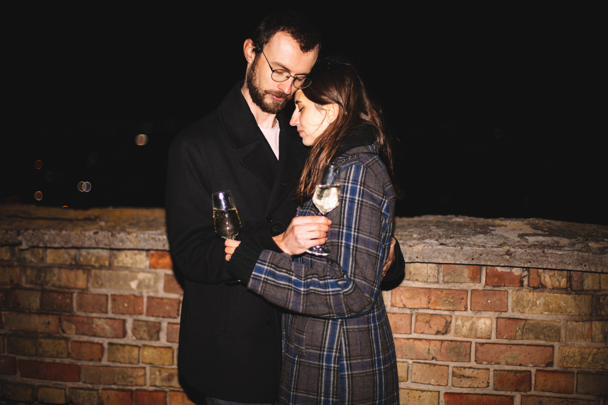 A man and woman in warm coats stand close together, holding glasses of white wine, looking into each other's eyes, against a brick wall at night