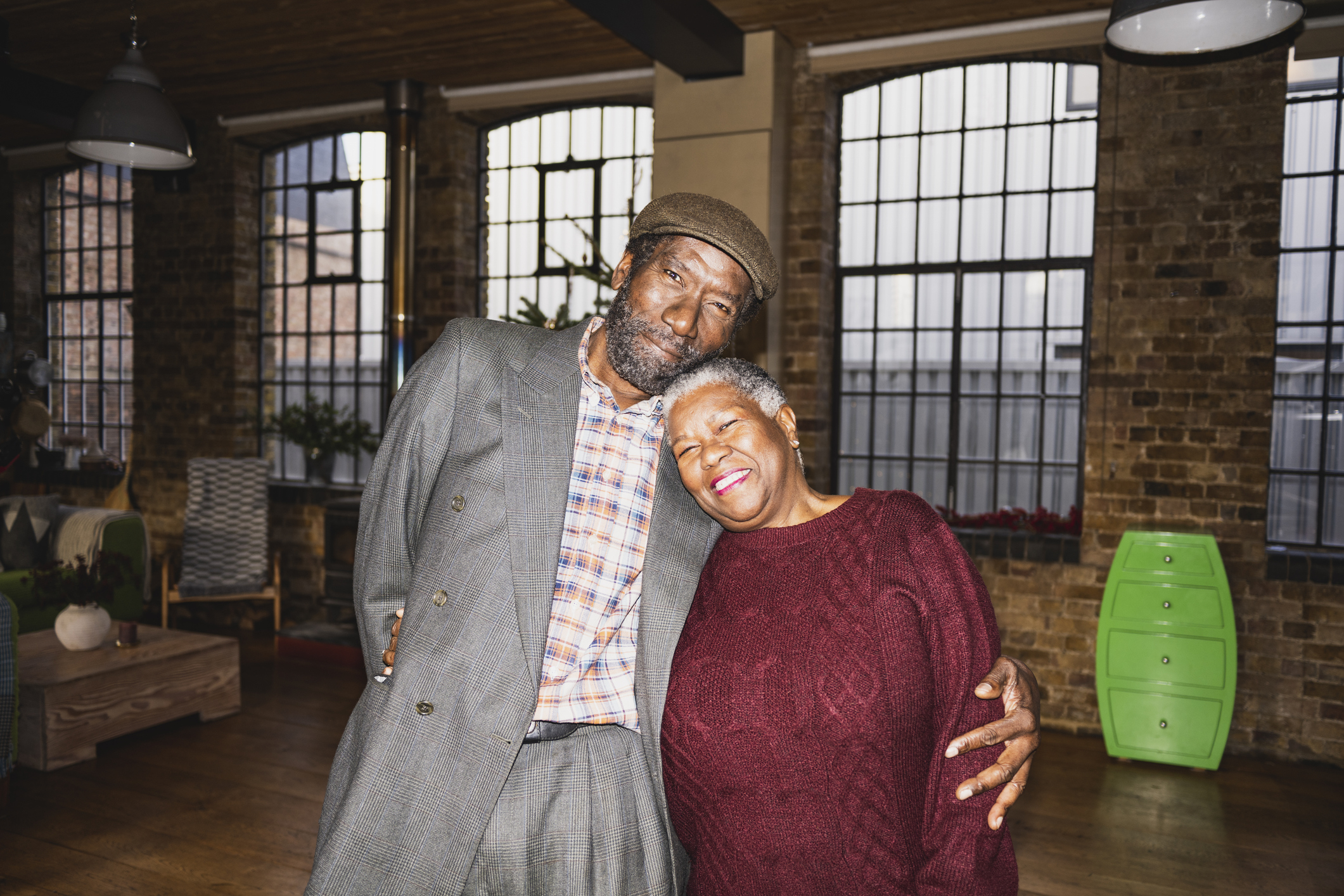 An elderly man in a gray suit and a woman in a burgundy sweater stand closely together, smiling warmly in a cozy room with large windows