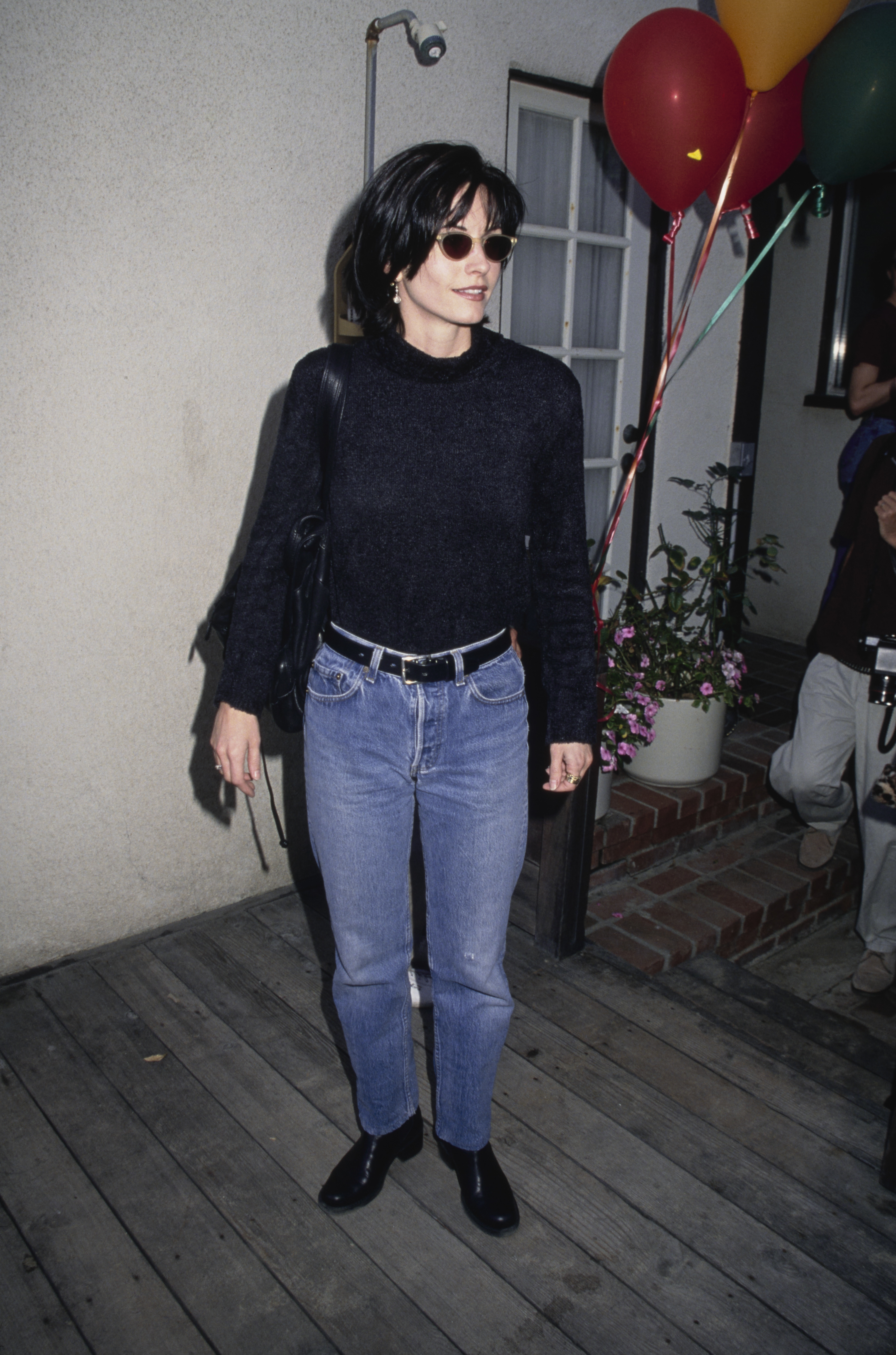 Courteney Cox standing on a wooden deck wearing sunglasses, a black sweater, blue jeans, black shoes, and holding a black handbag, with balloons in the background
