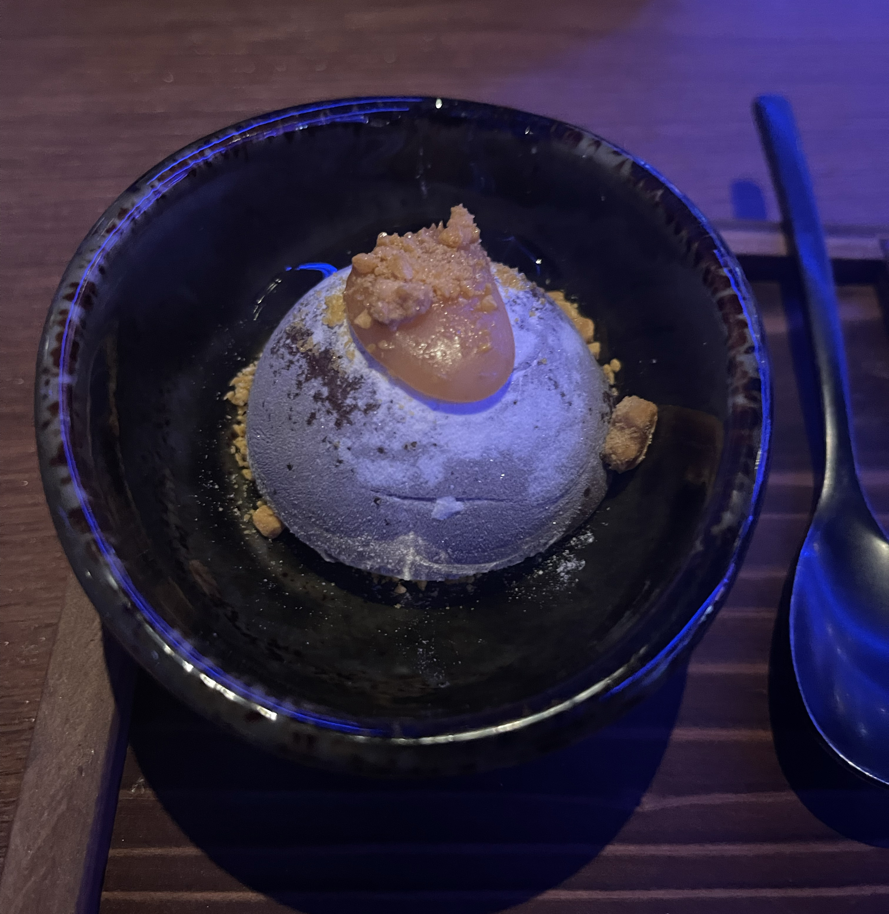 A plated dessert with a spherical dome, topped with crumbled bits and a sauce, accompanied by a spoon on a wooden tray