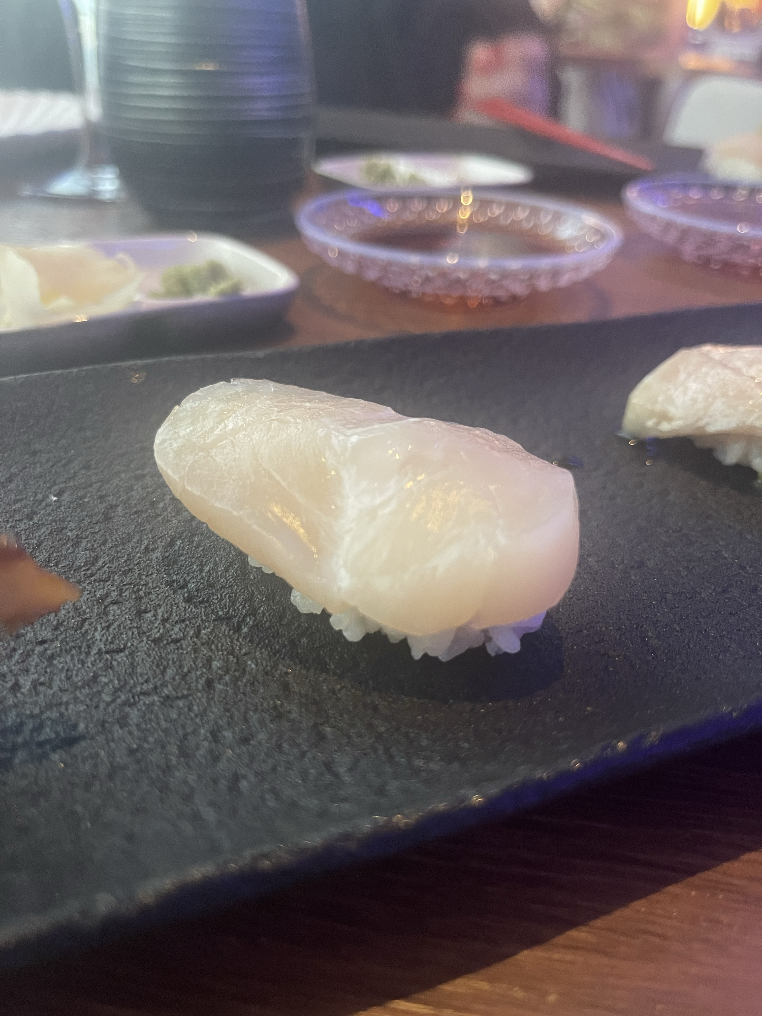 Close-up of a piece of sushi on a dark plate, with several small bowls holding sauces in the background