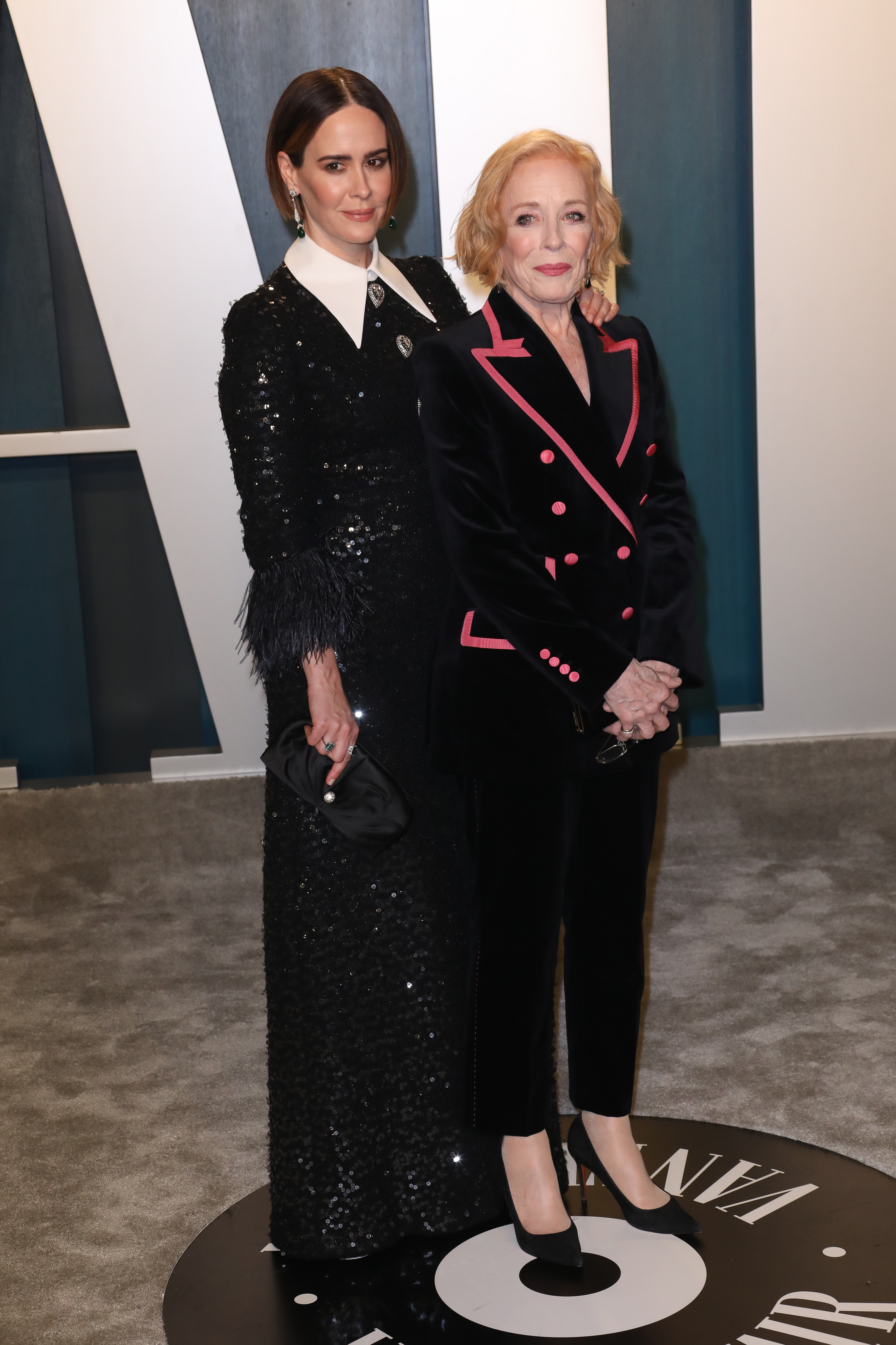 Sarah Paulson in a black sequined gown and Holland Taylor in a black suit with pink trim pose together on a red carpet event