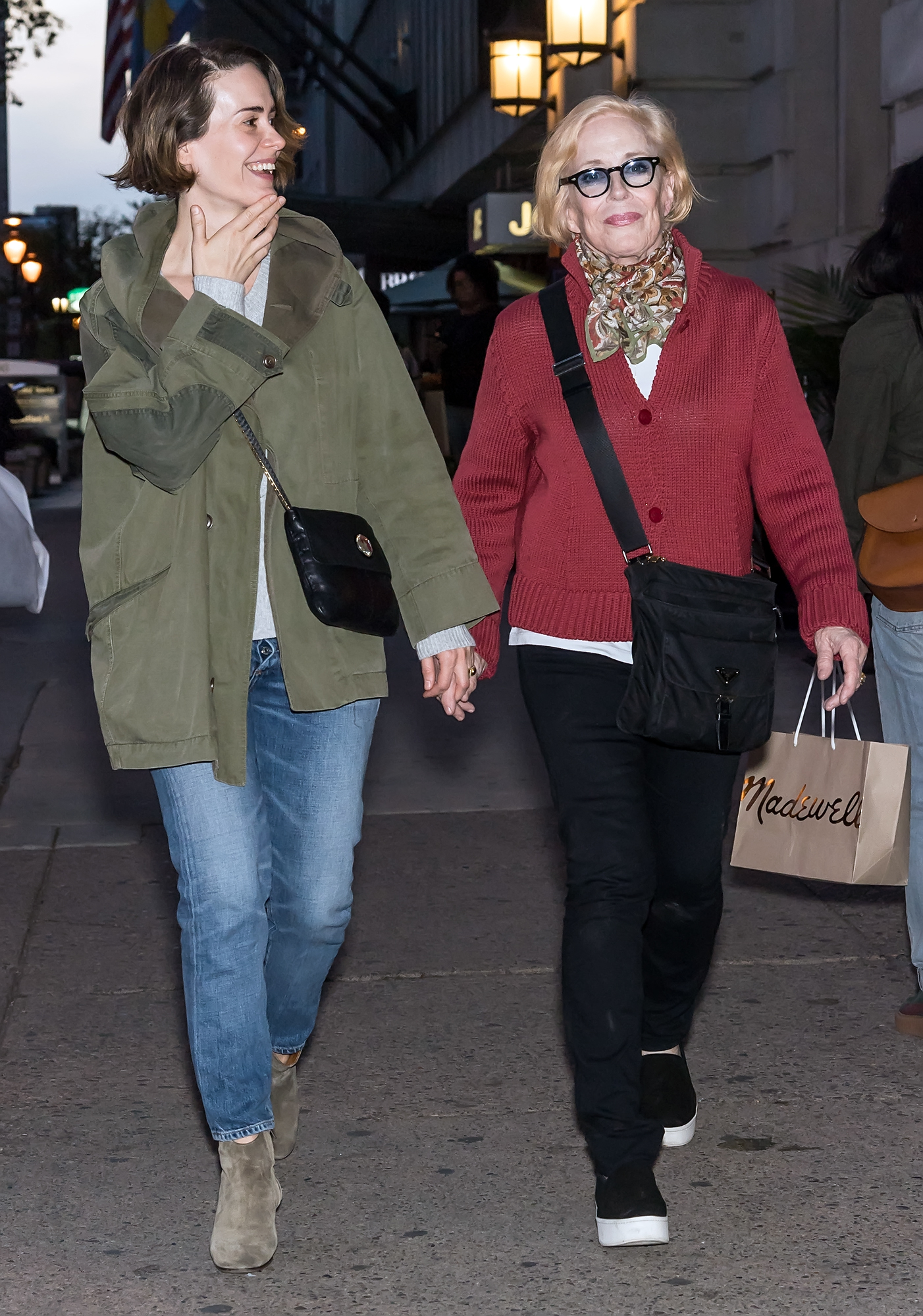 Sarah Paulson and Holland Taylor are walking down a city street holding hands. Holland carries a shopping bag and wears a red sweater. Both are smiling