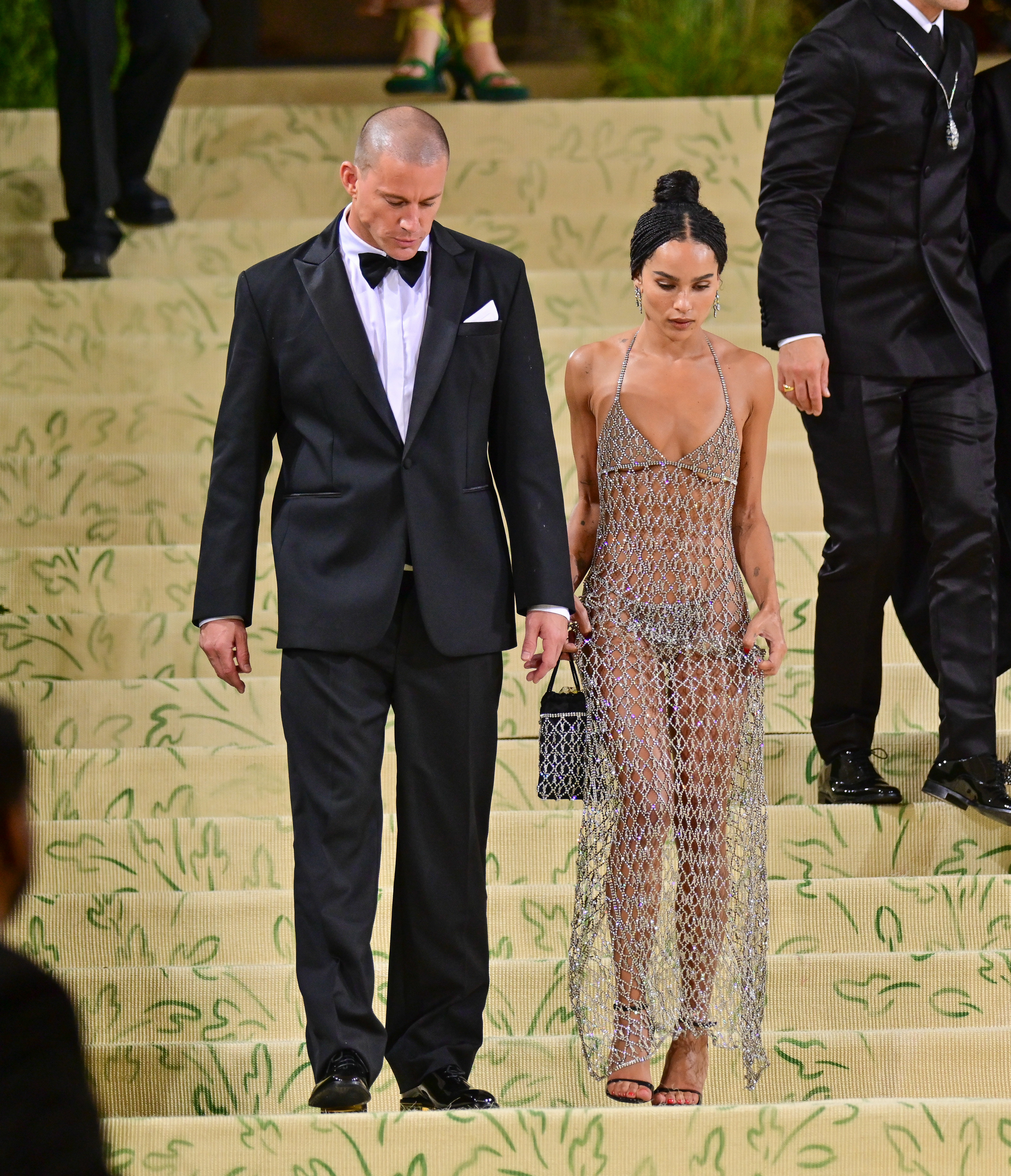 Channing Tatum in a black tuxedo and Zoë Kravitz in a sheer chainmail dress walk down stairs at a formal event