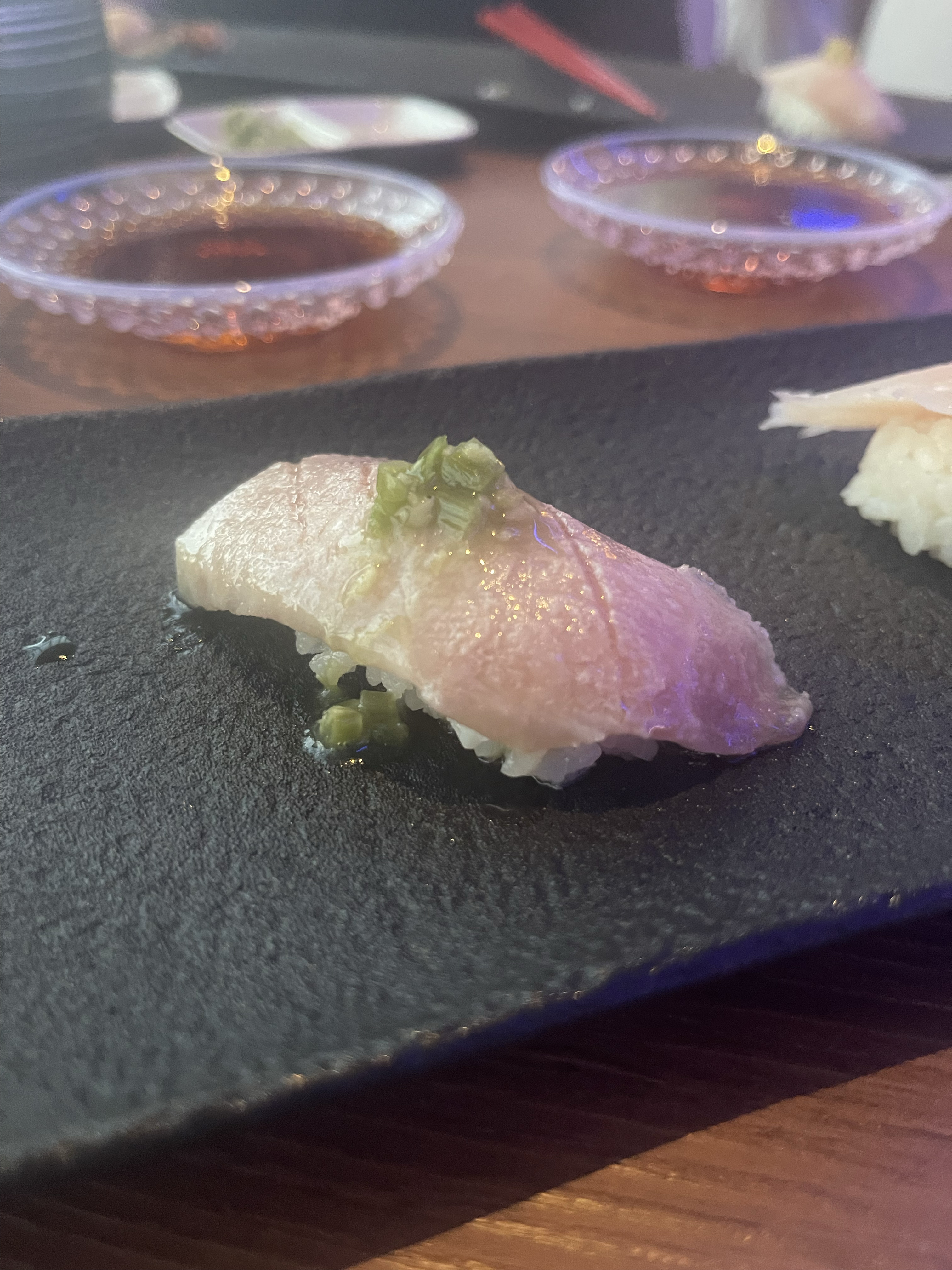 A piece of sushi with a topping of finely chopped green garnish on a black plate, with small saucers of dipping sauce in the background