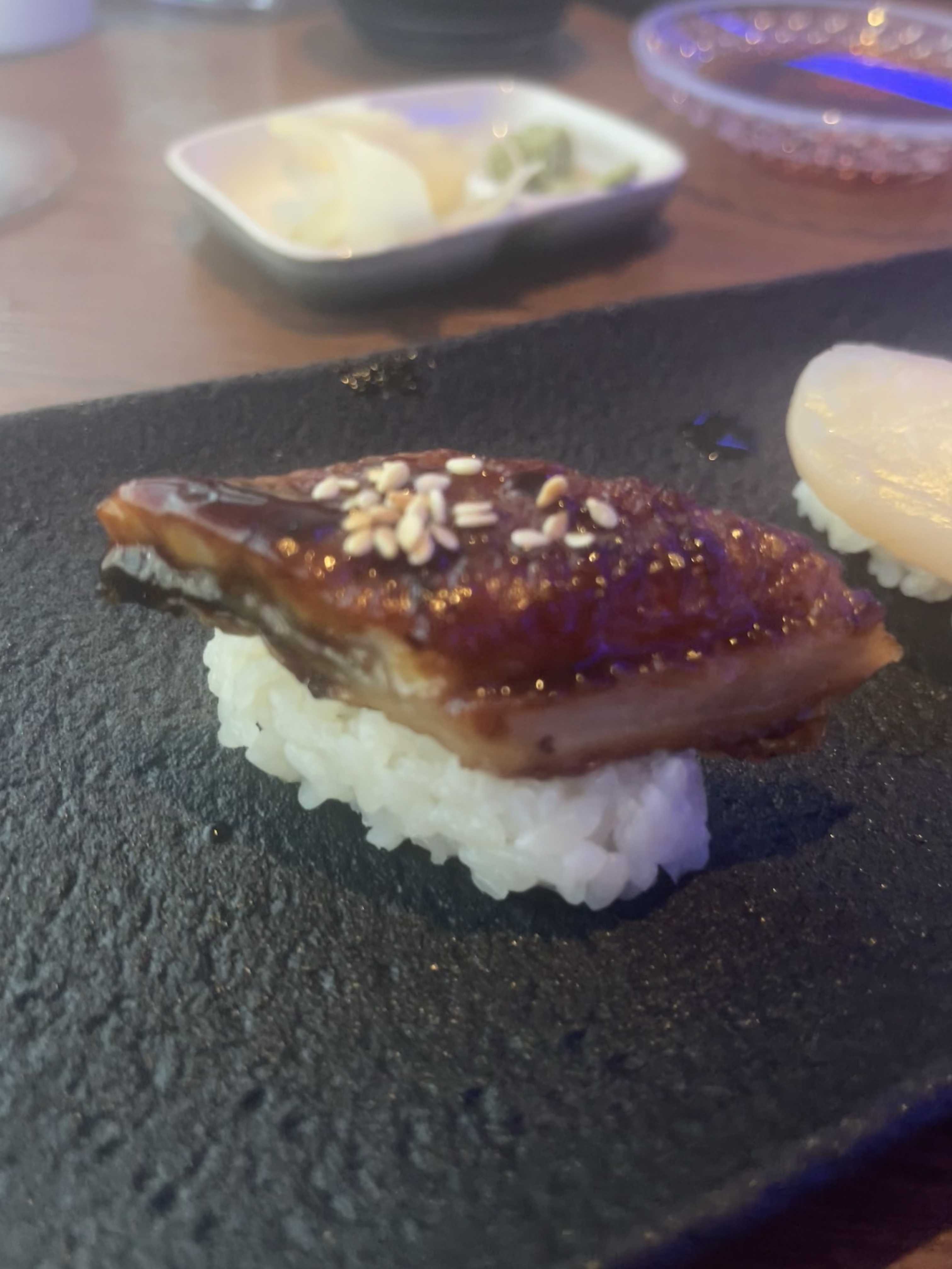 Close-up of a piece of sushi with a slice of eel and sesame seeds on top of rice, set on a dark plate. In the background, there are small dishes