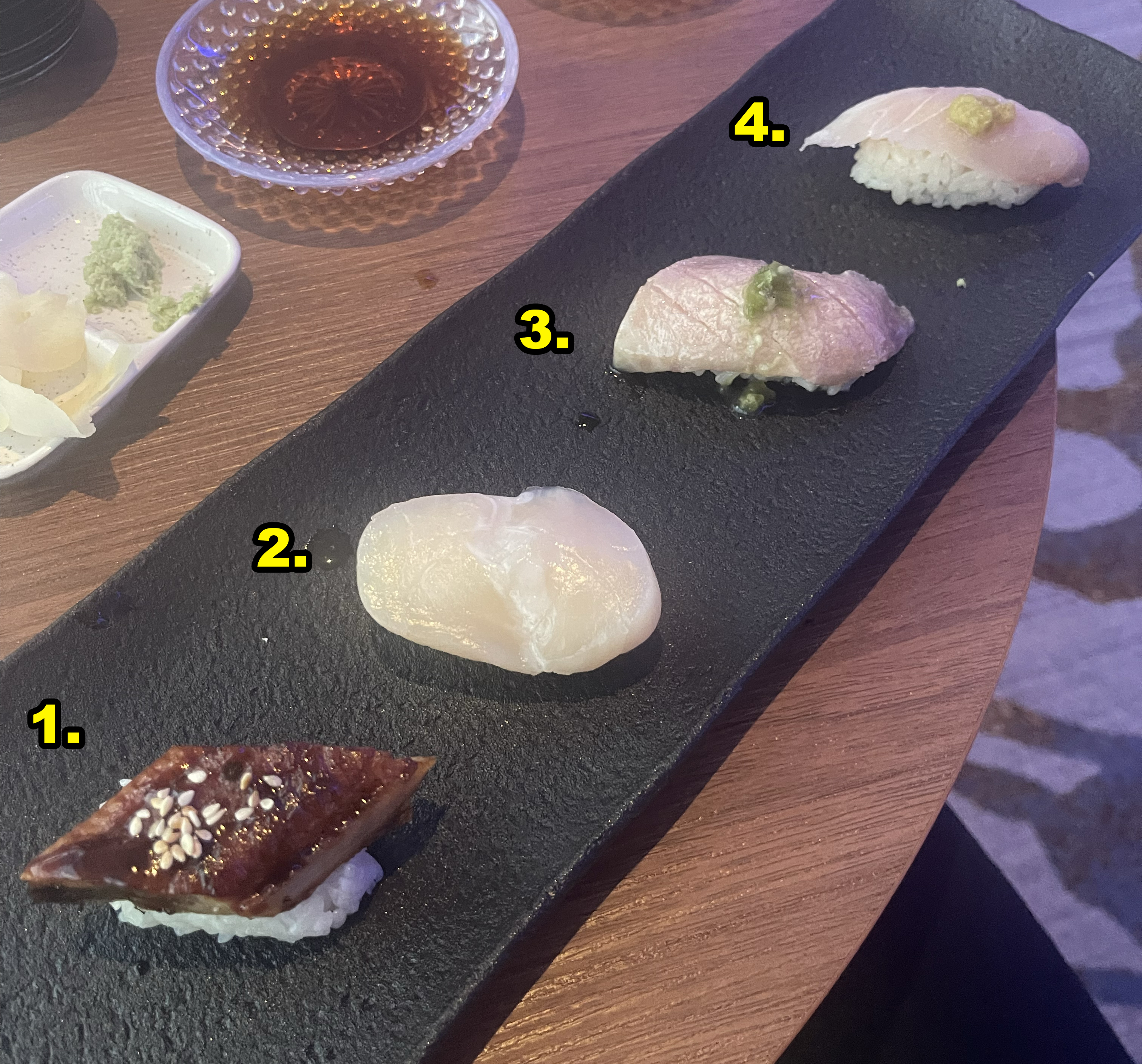 A close-up image of various sushi pieces numbered on a black rectangular plate, accompanied by soy sauce and wasabi on a wooden table