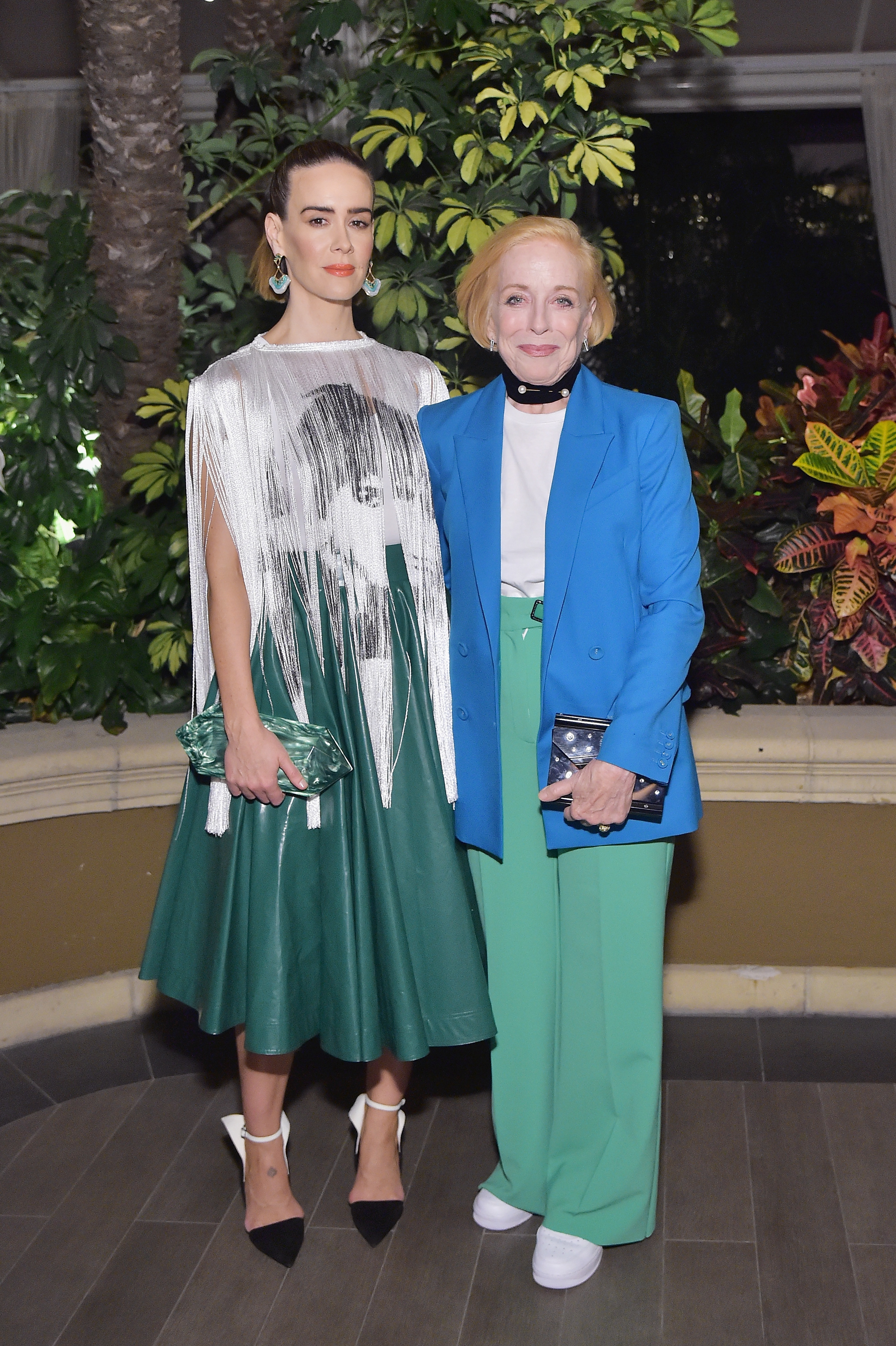 Sarah Paulson and Holland Taylor pose together, Sarah in a green skirt with a fringed top and high heels, and Holland in a blazer and pants