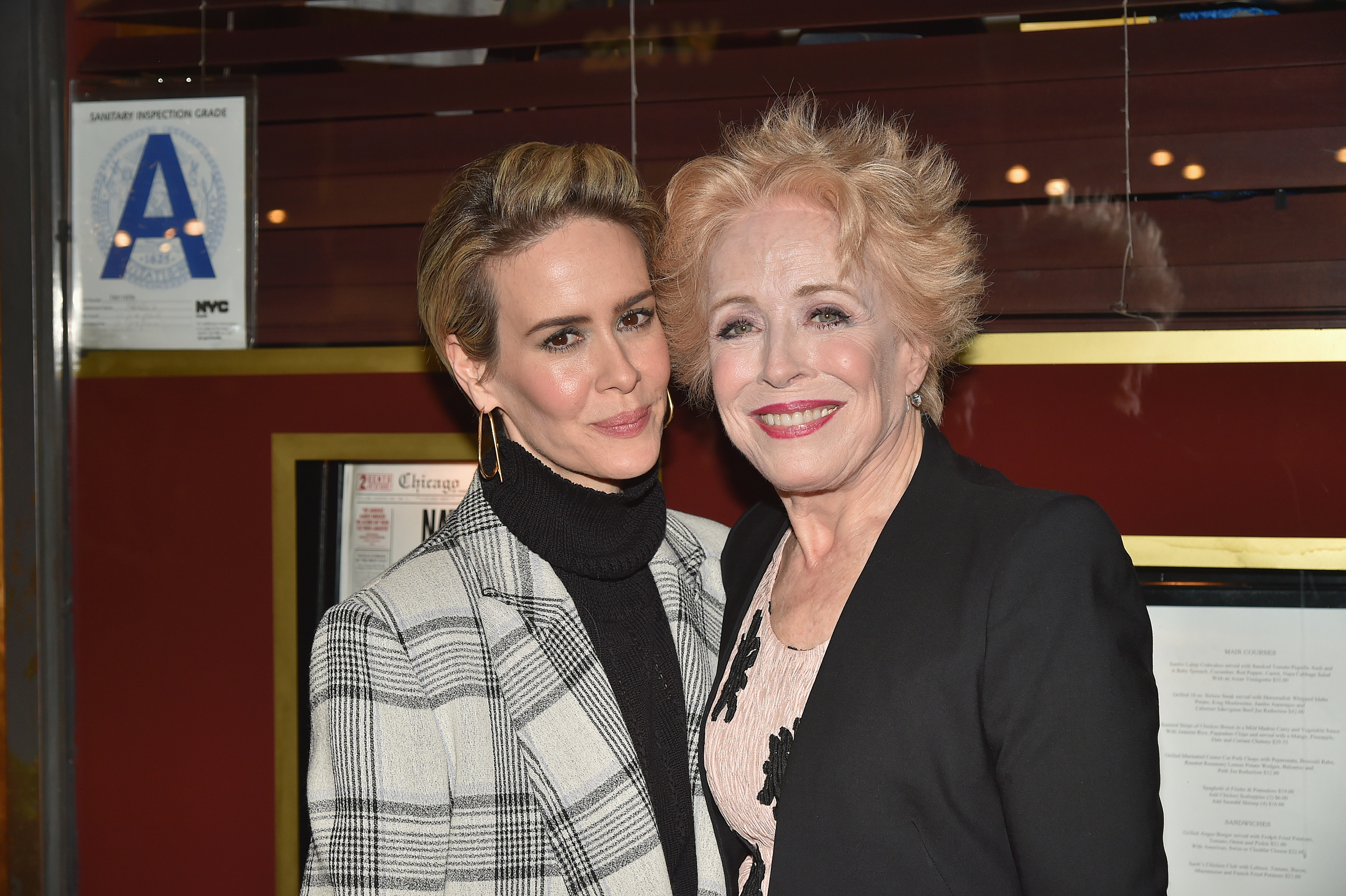 Sarah Paulson and Holland Taylor smile together at an event. Sarah wears a checkered coat over a turtleneck, and Holland sports a light-colored outfit