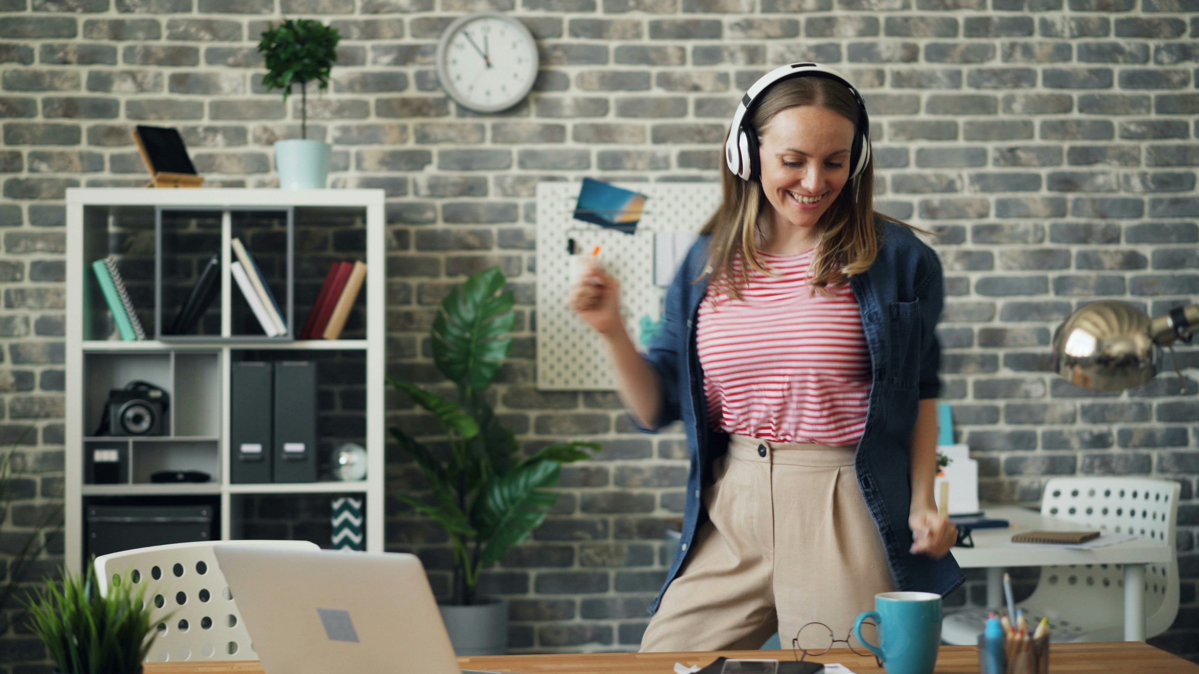 Woman in a casual office setting happily dancing with headphones on, in front of her workspace which includes a clock and shelves