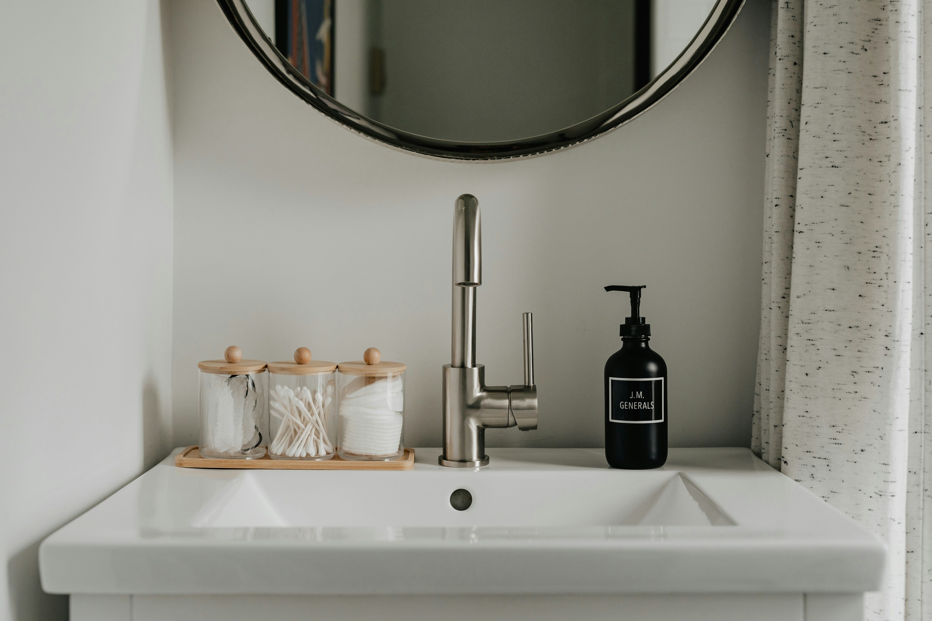 Modern bathroom sink with a circular mirror, cotton swabs, cotton pads in containers, and a black soap dispenser labeled "Pure Castile Soap."