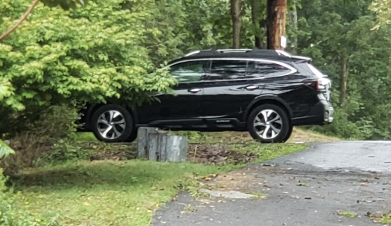 A black SUV parked on a grassy area near a forest, beside an unpaved road with trees in the background