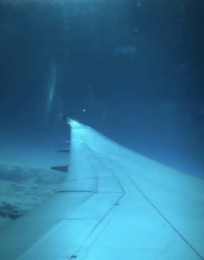 View from an airplane window showing the aircraft's wing stretching out over a layer of clouds and blue sky above