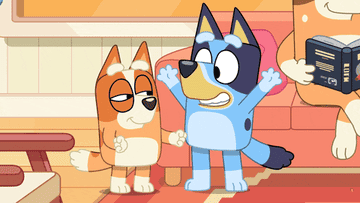 Bluey, an animated blue heeler puppy, stands excitedly beside Bingo, her orange heeler puppy sister, while an adult figure reads a book in the background