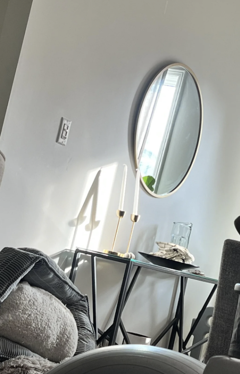 A round mirror hangs on a wall above a small table with two white candles, a clear jar, and some books. A pile of blankets is seen on a chair to the left