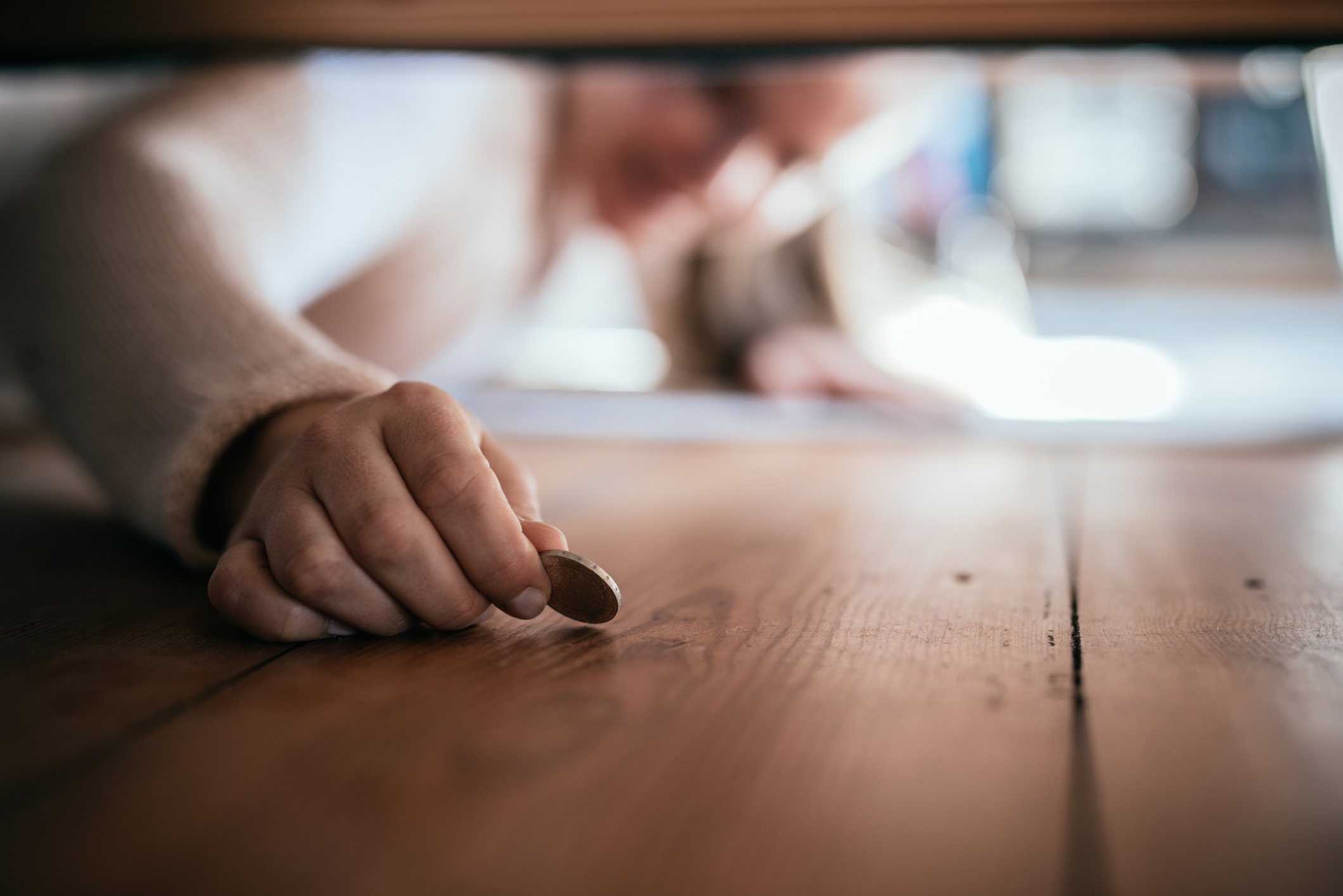 Person reaching under furniture to pick up a small coin from a wooden floor, face blurred and not fully visible in the background