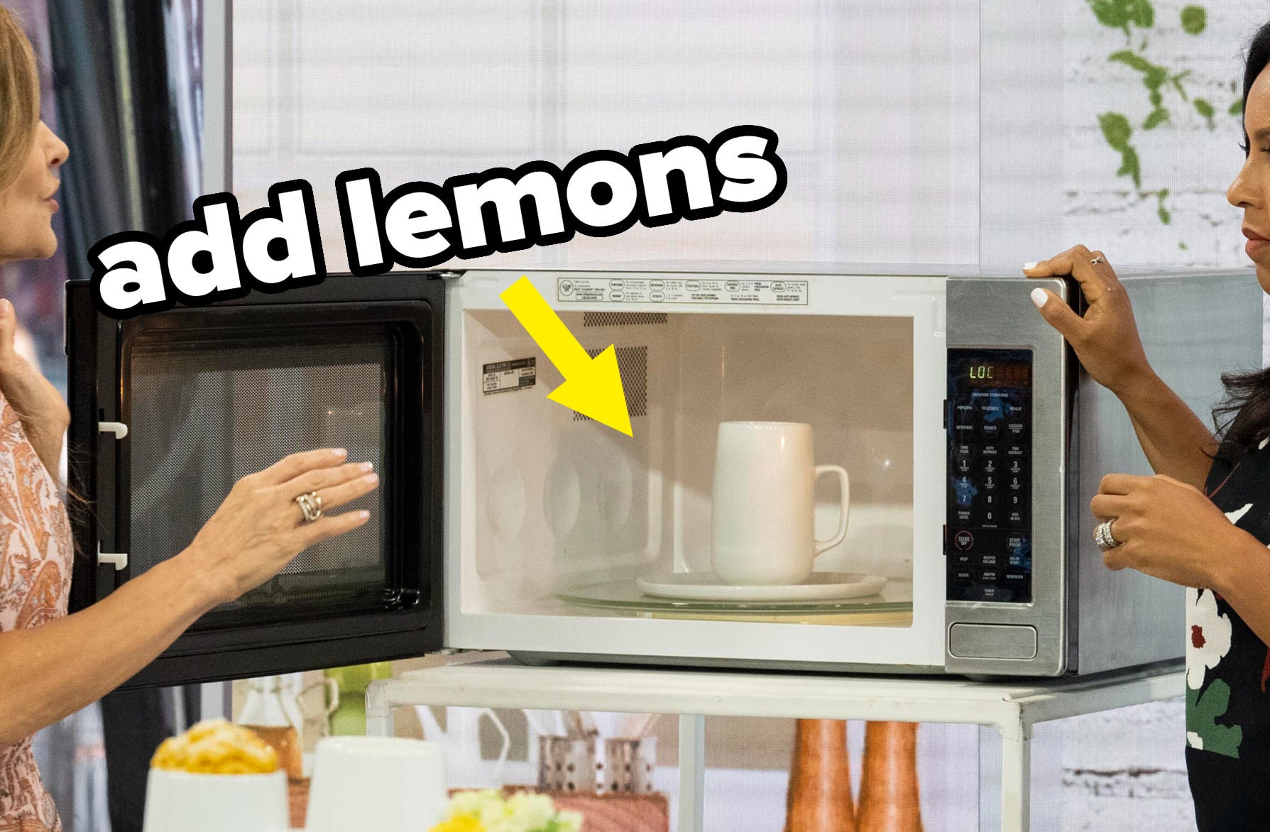 Two women stand at a kitchen counter. One shows a microwave oven with a mug inside to the other. Various kitchen items are on the counter around them