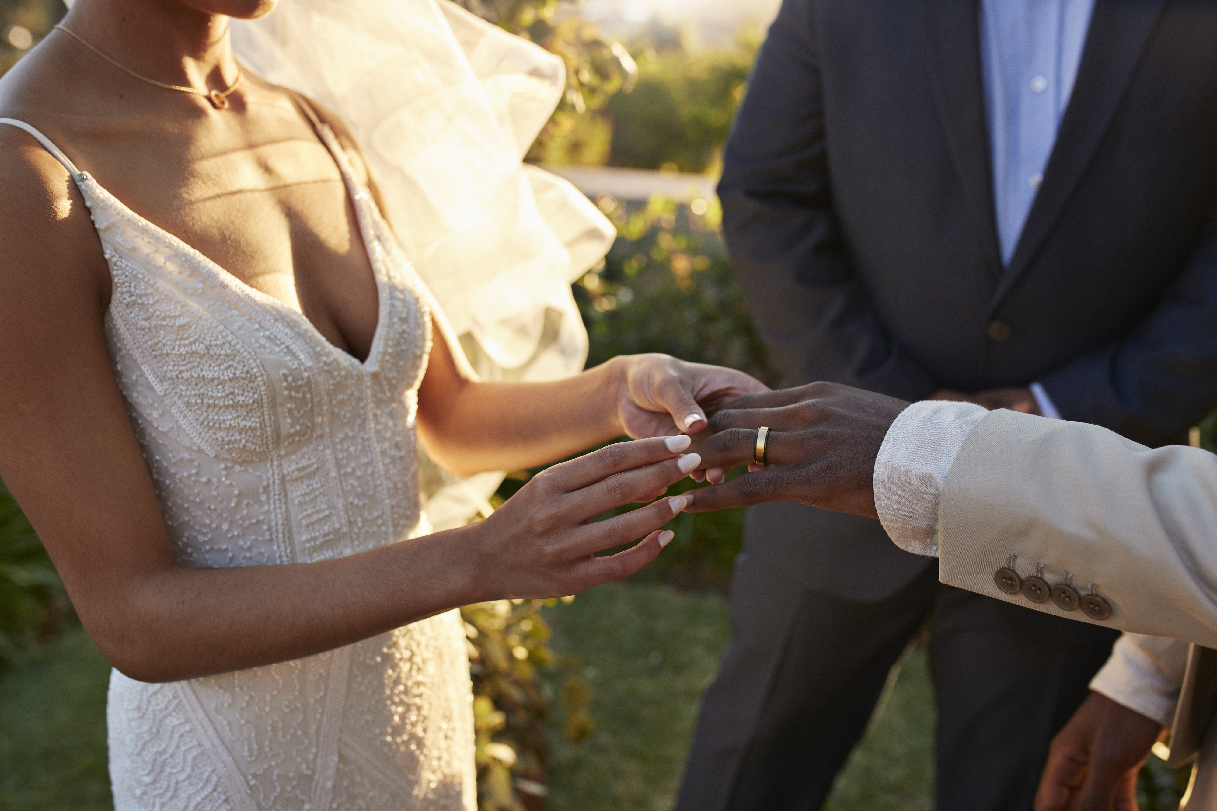 A bride in a sleeveless, beaded wedding gown places a ring on the groom's finger during an outdoor ceremony officiated by a man in a suit