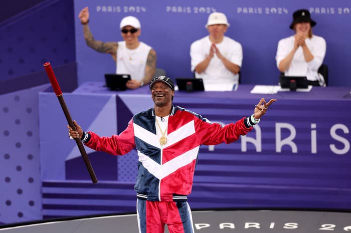 Snoop Dogg holds a baton and smiles in a tracksuit, in front of judges at the Paris 2024 event. Three judges clap behind him