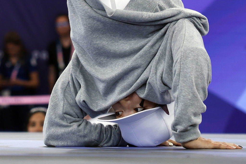 Person breakdancing, wearing a gray hoodie and a white cap, performing a headstand on a stage during a competition