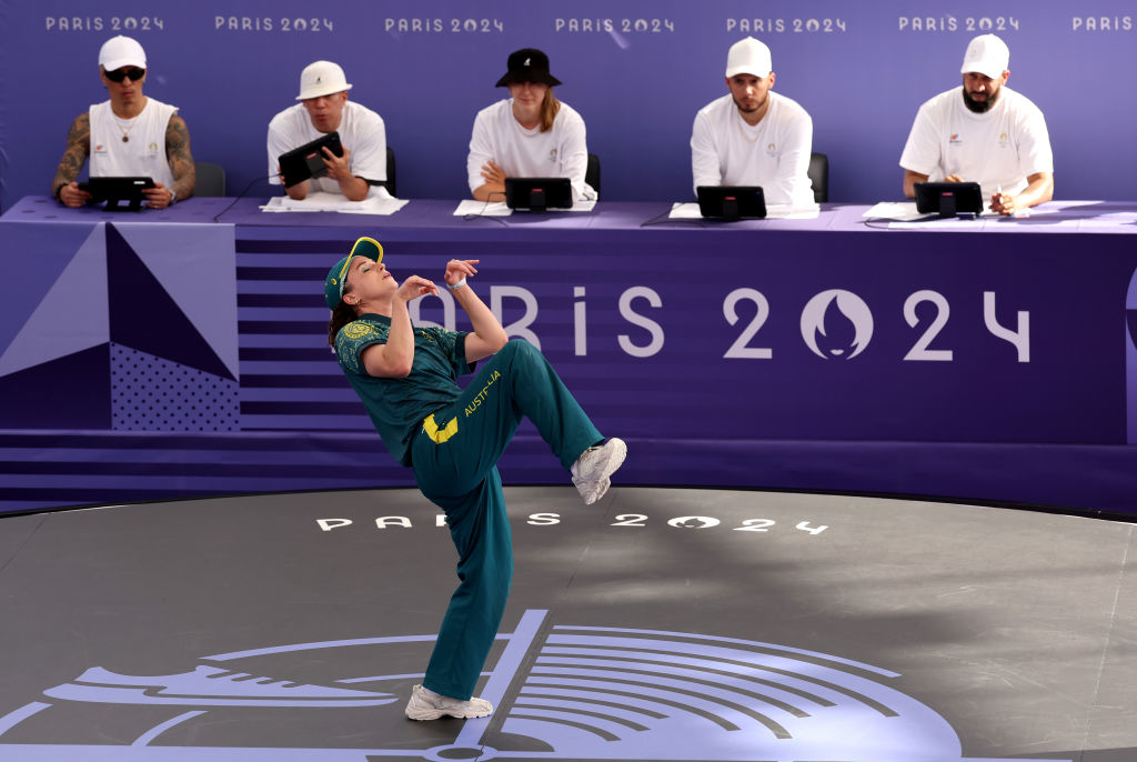 Raygun breakdancing in front of judges at Paris 2024 event, wearing sports attire and posing like a velociraptor. Five judges, seated with tablets, watch attentively
