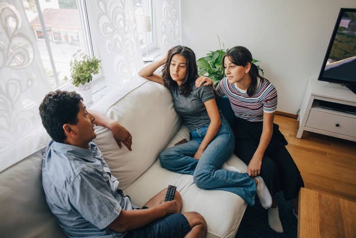 Three people, not identified, sitting in a living room. A man holds a remote while two women sit close together on the couch, appearing to have a conversation