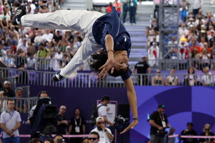 Breaking athlete performs a mid-air flip at a sporting event. Spectators and media can be seen in the background