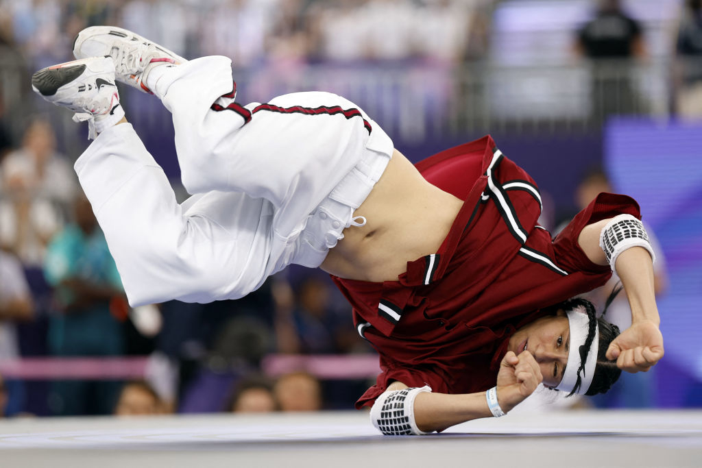Anti in athletic clothing performs an impressive breakdancing move, balancing on one arm and with their legs in the air