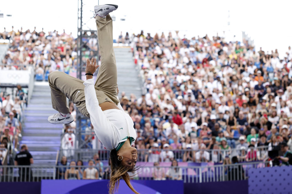 B-Girl performs a mid-air flip during the breaking competition at the 2020 Tokyo Olympics, with a large crowd in the background