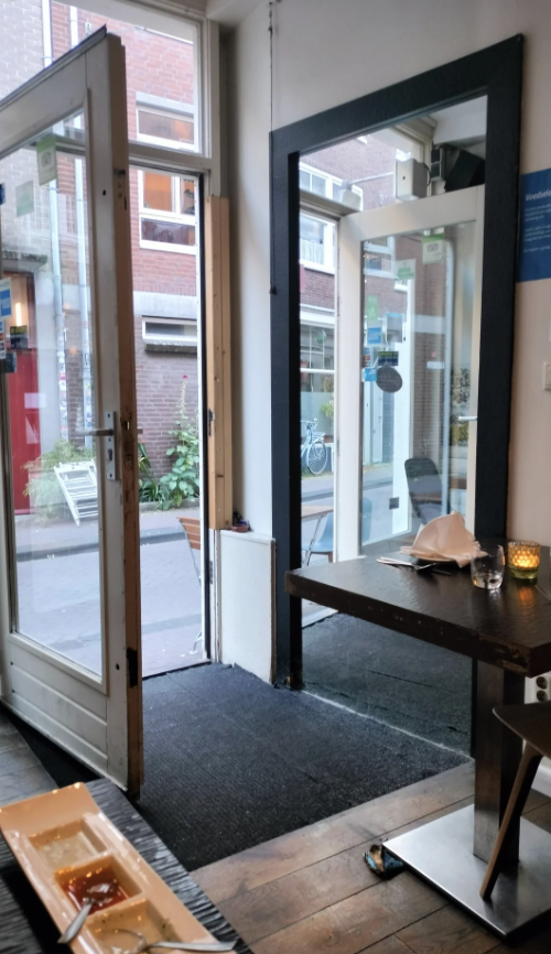 Open door leading to a street outside a small cafe, with a table, chairs, and a candle on the table inside. A bicycle is visible through the door