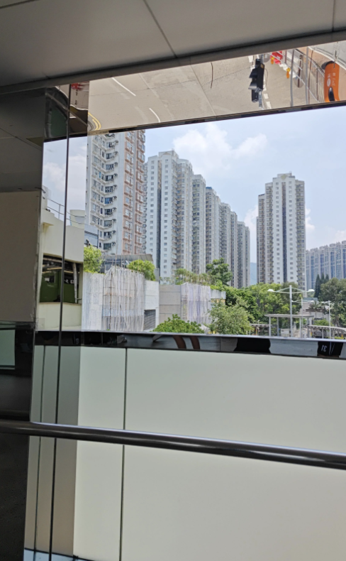 View of tall residential buildings through a glass window with reflections of the room's interior. Greenery and other buildings are visible in the background