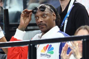 Snoop Dogg is seen wearing a jacket with an NBC logo and the text "SnoopDogg," holding sunglasses while making a surprised expression