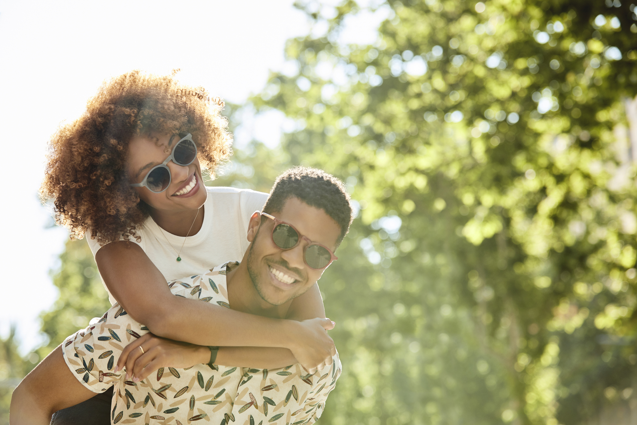 A person gives their partner a piggyback ride outdoors, both smiling and wearing sunglasses. The background includes trees and a bright, sunny ambiance