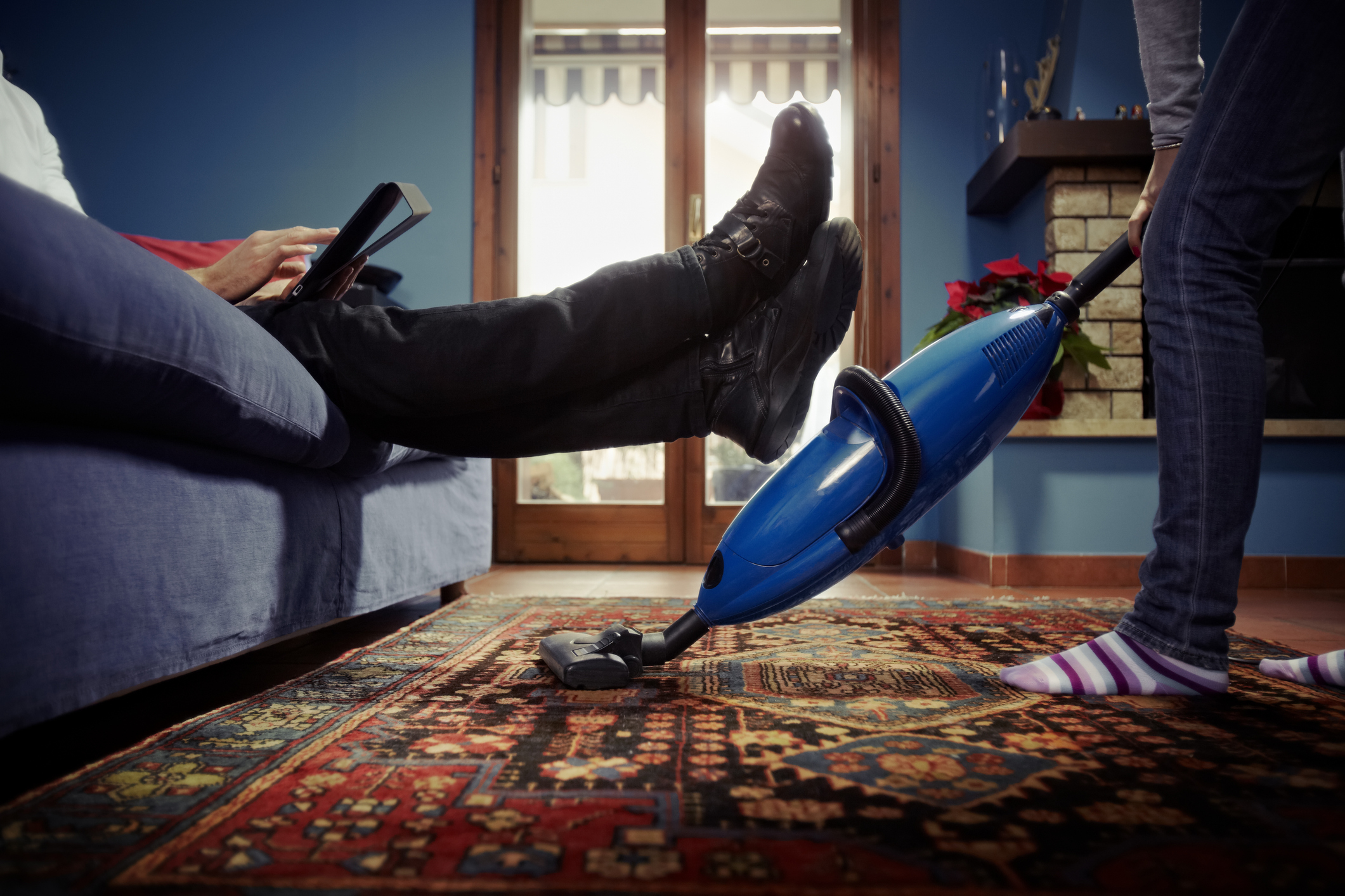 A person uses a vacuum cleaner on a patterned rug while another sits on a couch with their feet up, using a tablet