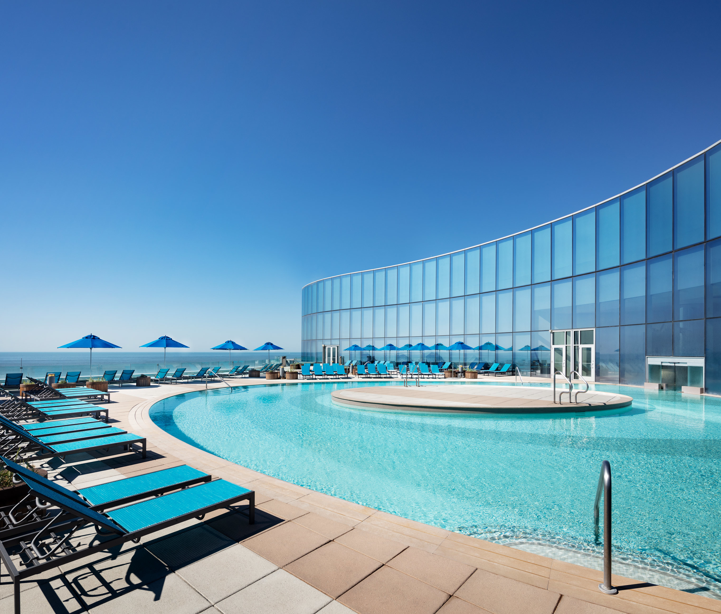 An outdoor pool area with lounge chairs and umbrellas beside a building with large glass windows, reflecting a spacious and vibrant atmosphere