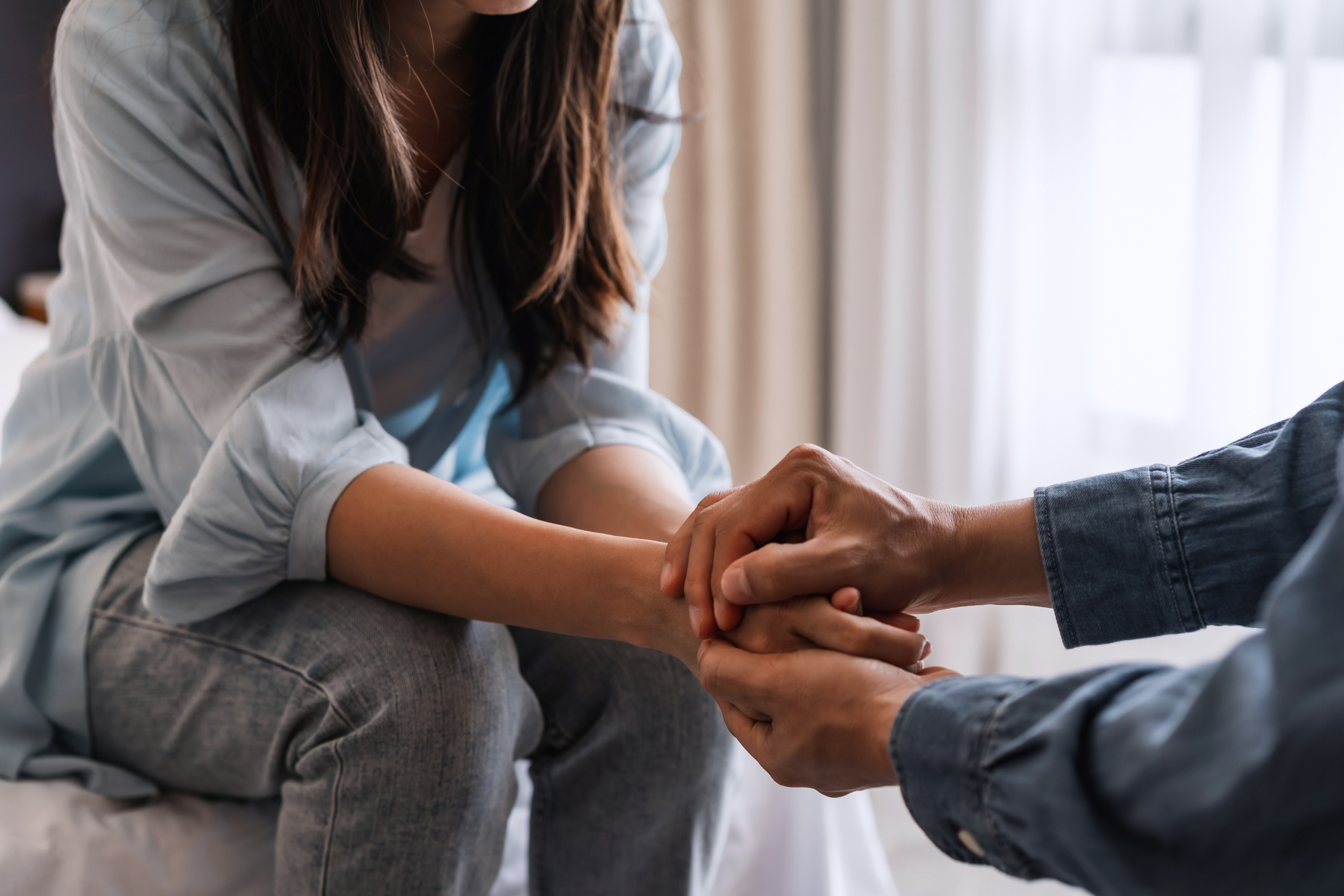 A couple sits closely on a bed; she holds his hands in hers. The moment appears intimate and heartfelt