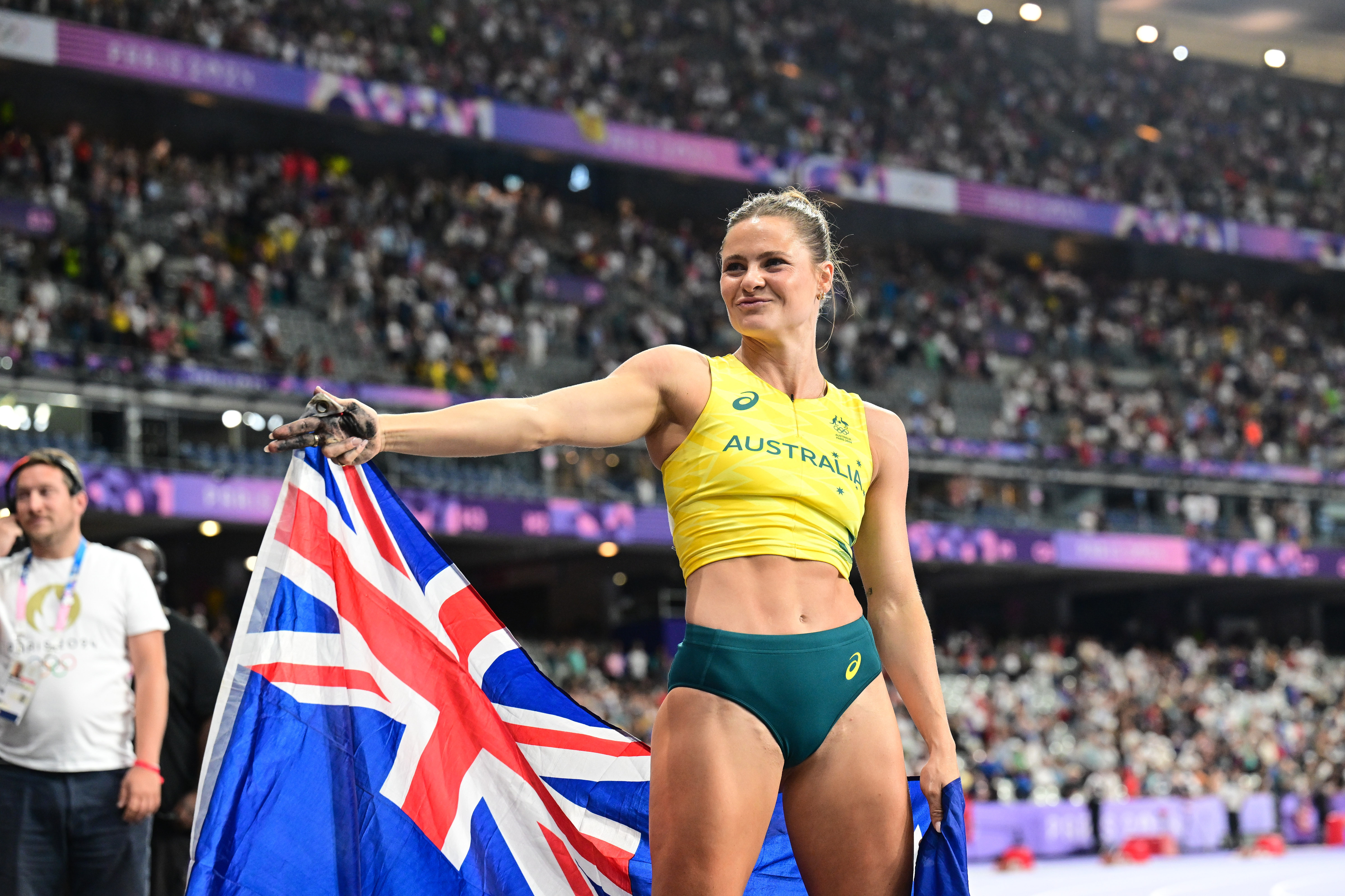 An athlete, wearing a yellow top and green shorts with &quot;Australia&quot; printed on the top, proudly holds the Australian flag in a stadium filled with spectators