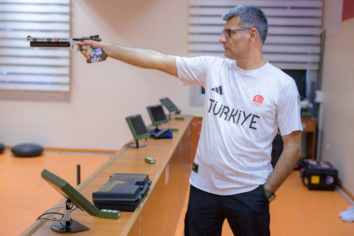 A man wearing glasses and a T-shirt with &quot;Türkiye&quot; and a crescent moon and star emblem is practicing target shooting indoors with a pistol