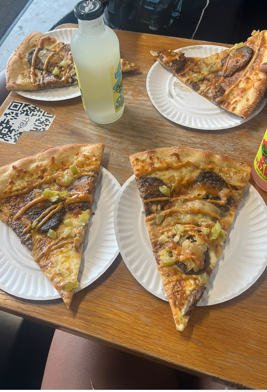 Several slices of pizza with various toppings on white paper plates are on a wooden table. A QR code is visible. There are drinks in plastic bottles