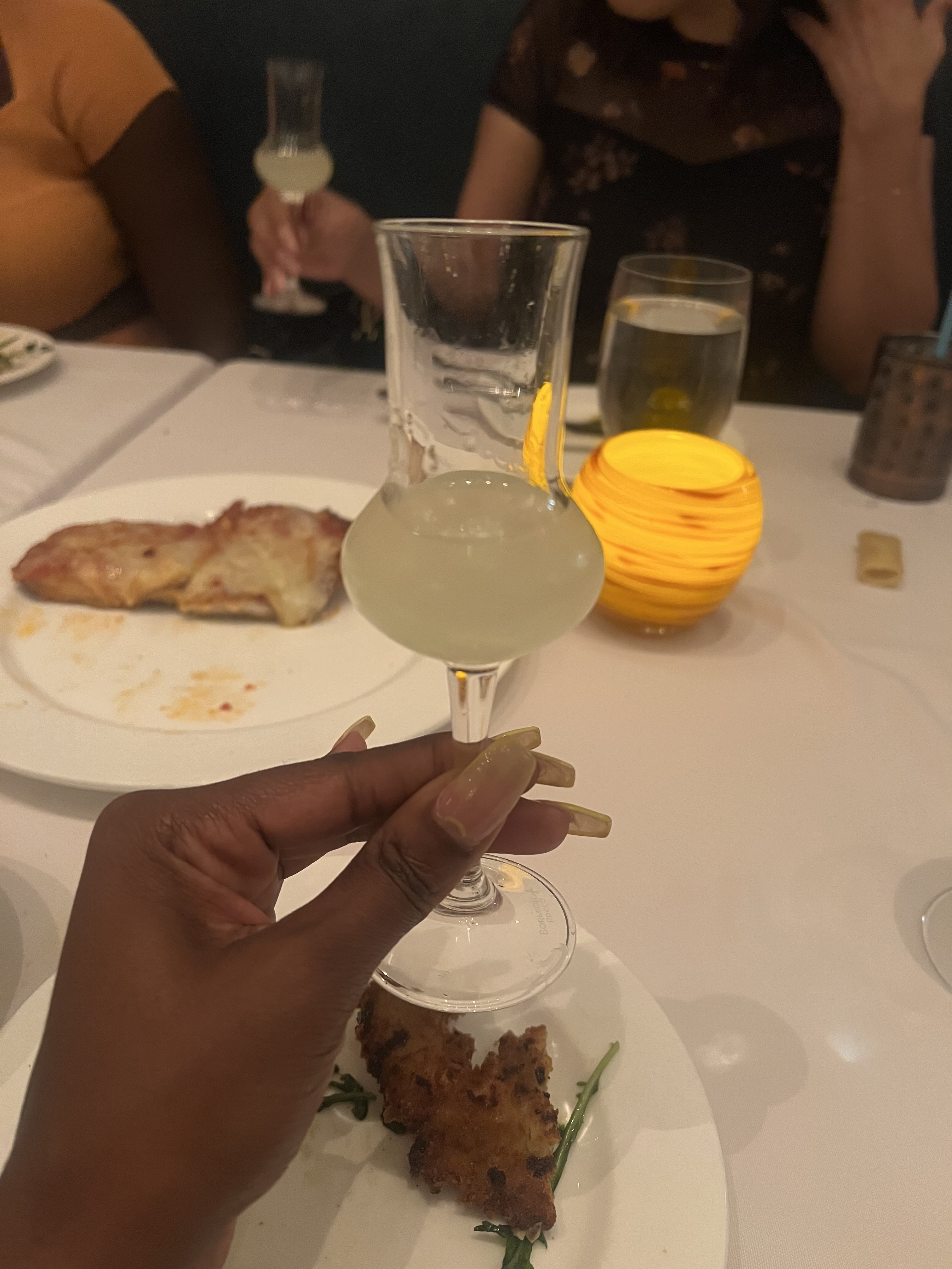A hand holding a glass of a pale yellow cocktail, with a table setting including a candle, plates with food, including a slice of pizza and a fried item