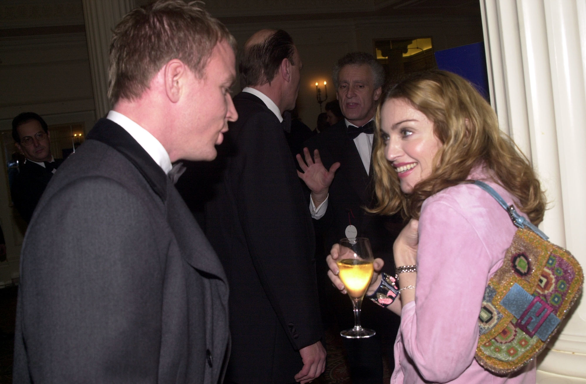 A man and a woman at a formal event. The woman is holding a drink and wearing a stylish outfit with a colorful bag, while the man is in formal attire. Names are unknown