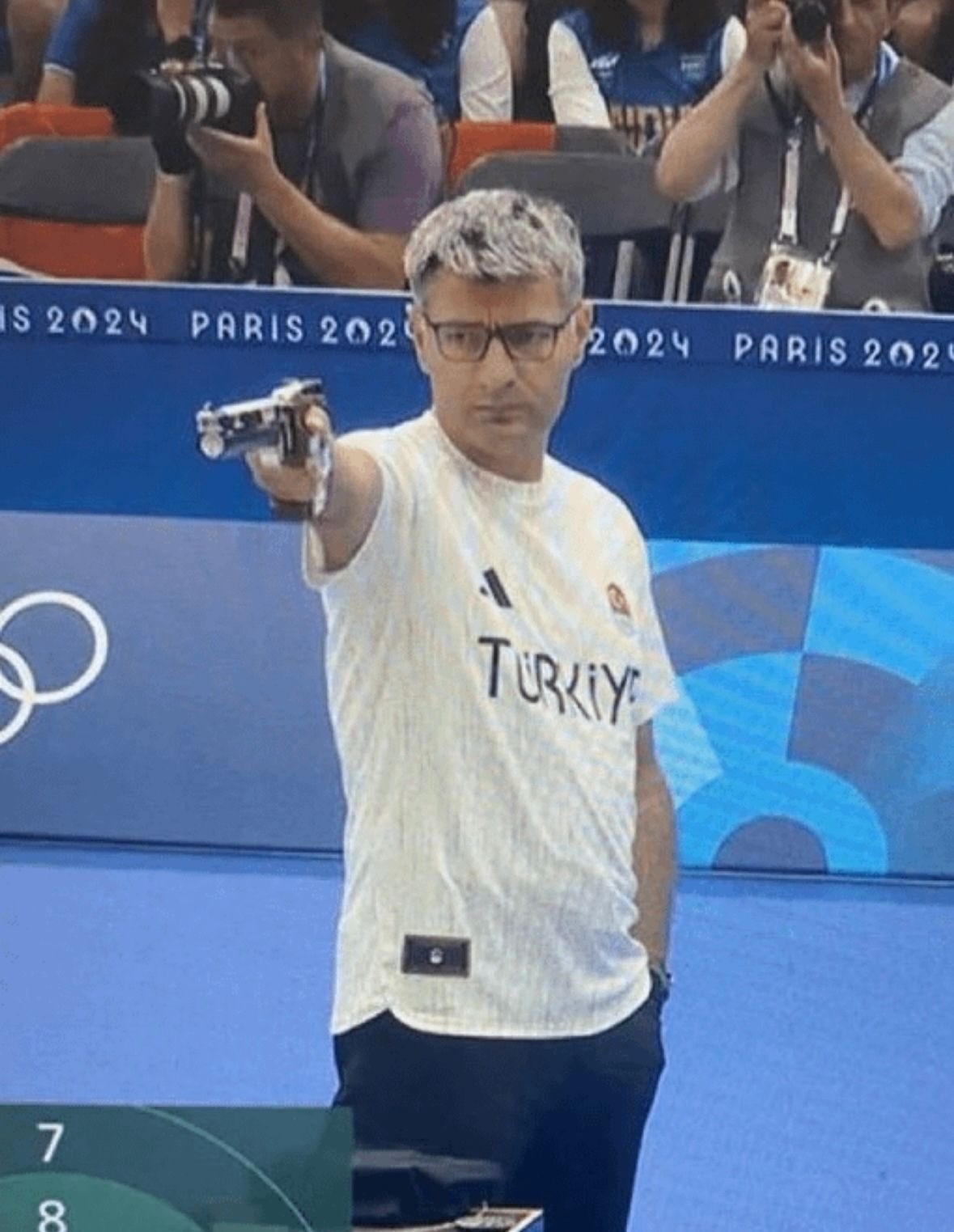 Male athlete wearing a &quot;Turkey&quot; labeled shirt, aiming a pistol at a sports event in the Paris 2024 Olympics. Photographers in the background