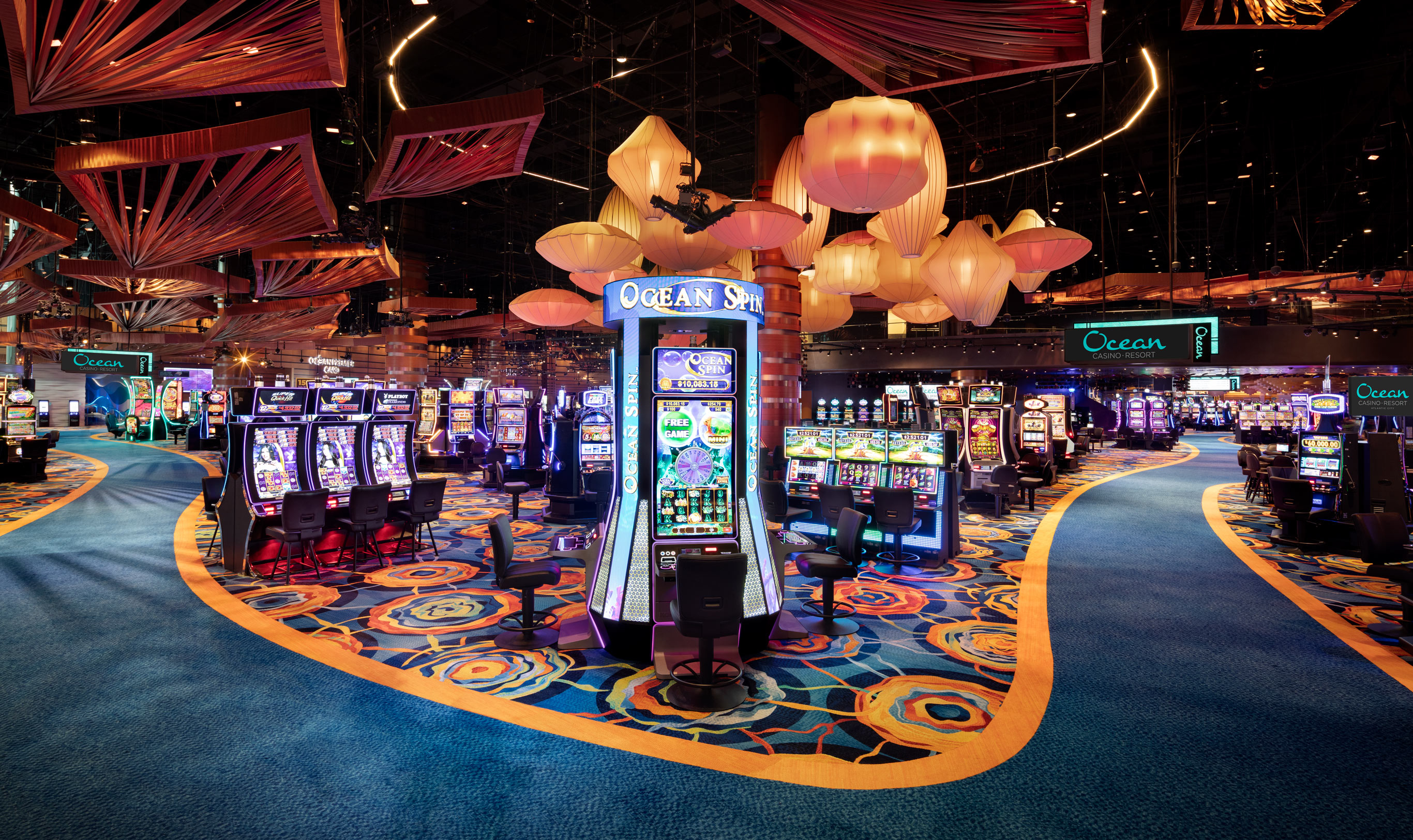 A spacious casino floor with rows of slot machines and intricate, colorful carpet patterns. Large ceiling decorations are prominently hung above