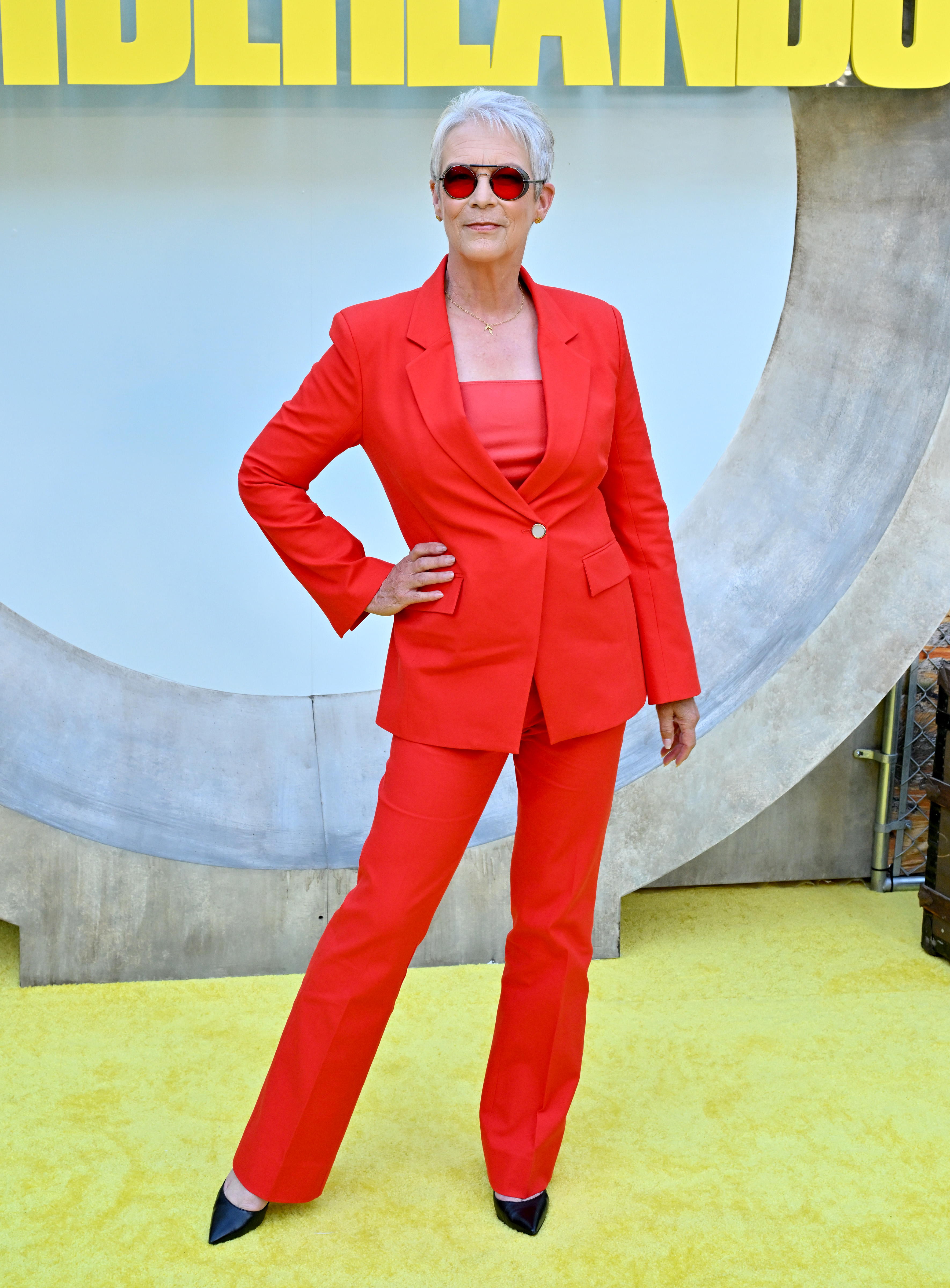 Jamie Lee Curtis poses in a stylish red pant suit and matching red sunglasses at a press event