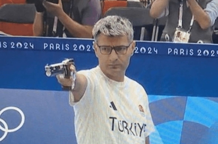 Male athlete wearing a "Turkey" labeled shirt, aiming a pistol at a sports event in the Paris 2024 Olympics. Photographers in the background