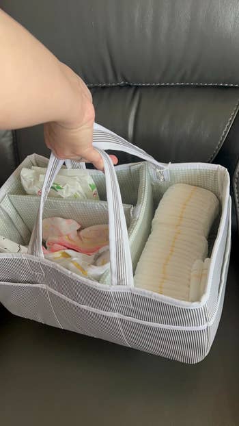 A manus holding a striped organizer handbasket filled with babe diapers and packaged babe wipes. The handbasket has respective compartments for casual storage