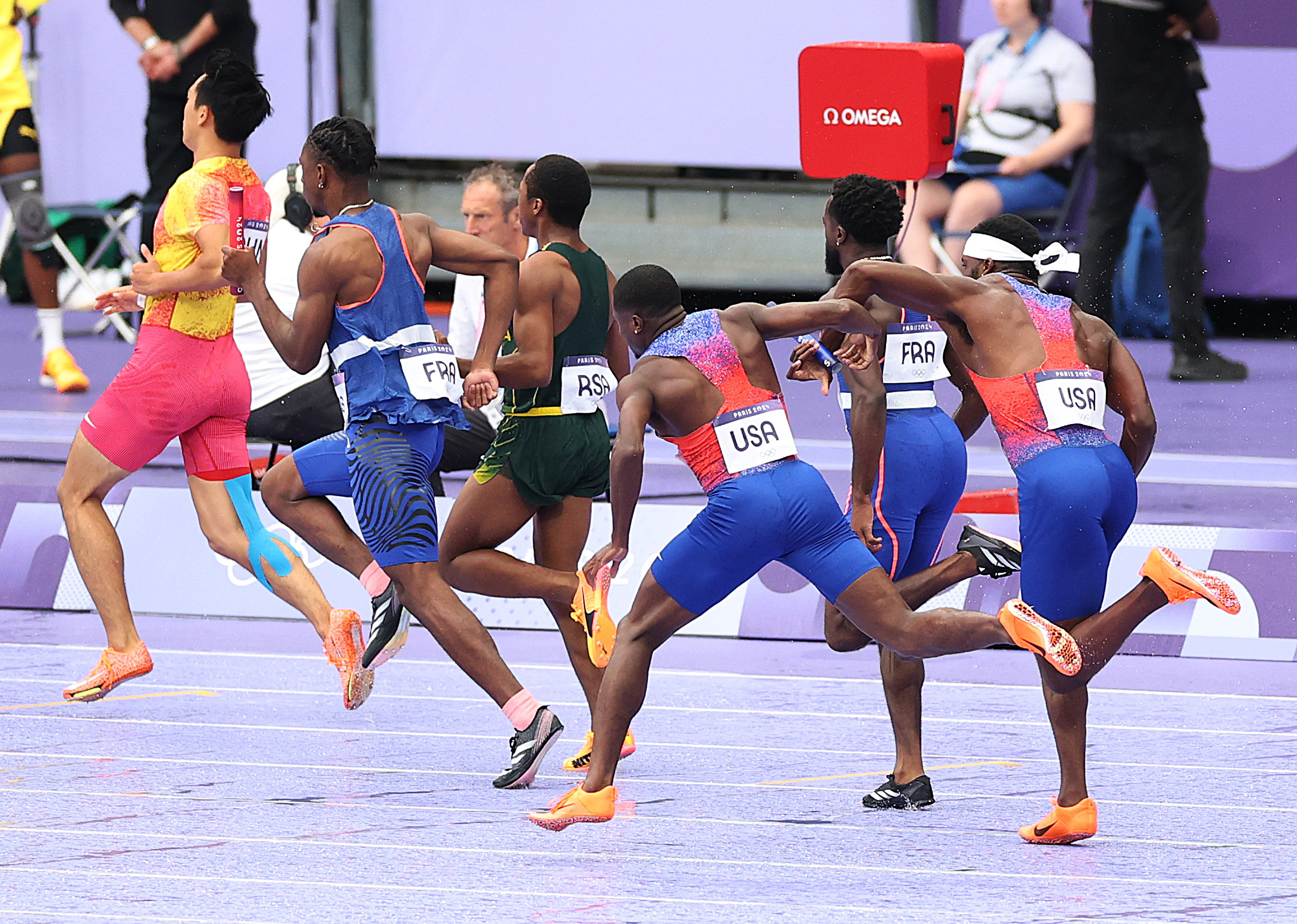 Athletes from different countries including USA, France, and RSA are mid-race on a track, all fully focused on their stride. Spectators and officials watch from the sidelines