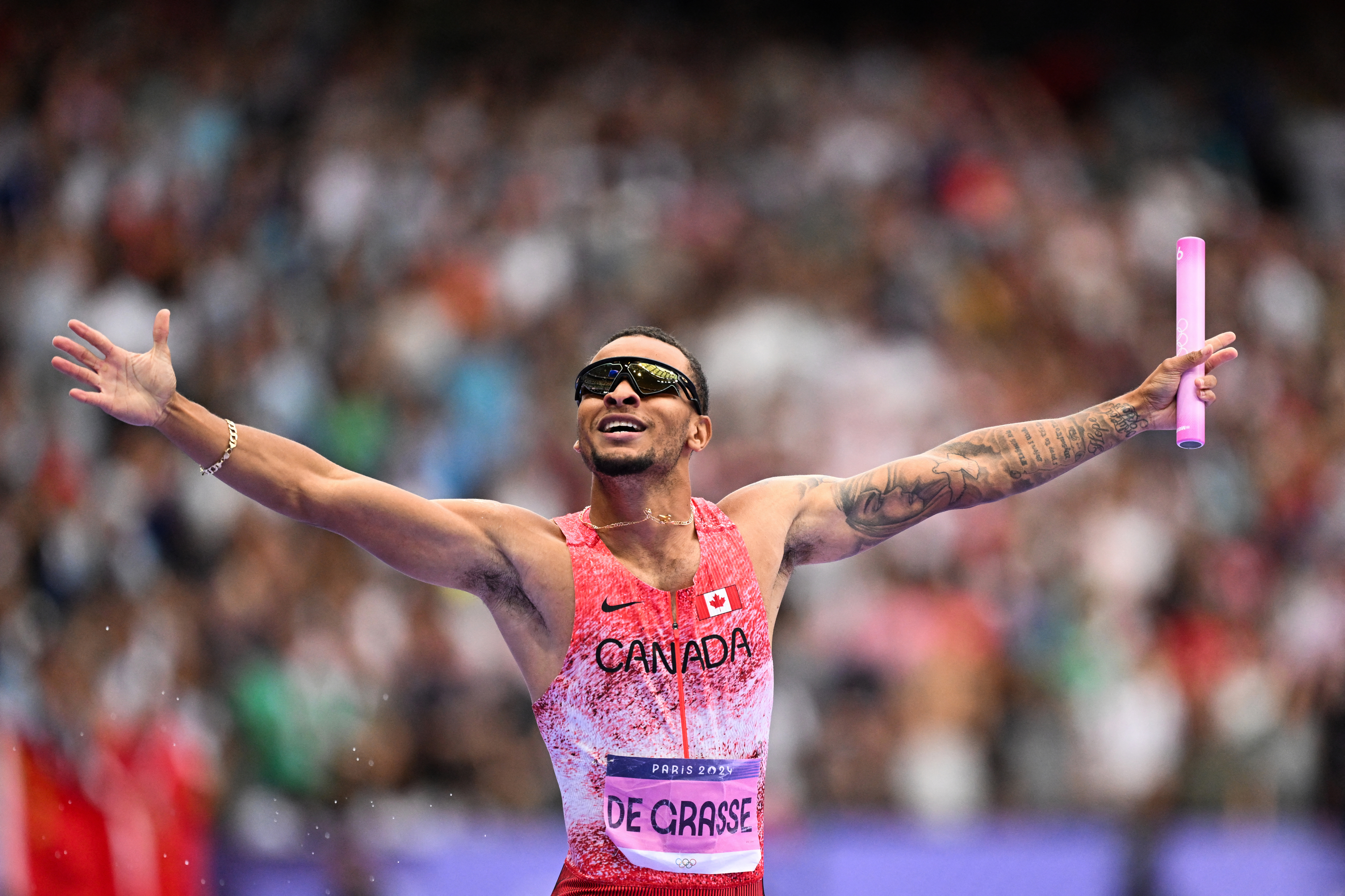Andre De Grasse, wearing a Canada jersey and sunglasses, spreads his arms in celebration while holding a baton at a track and field event