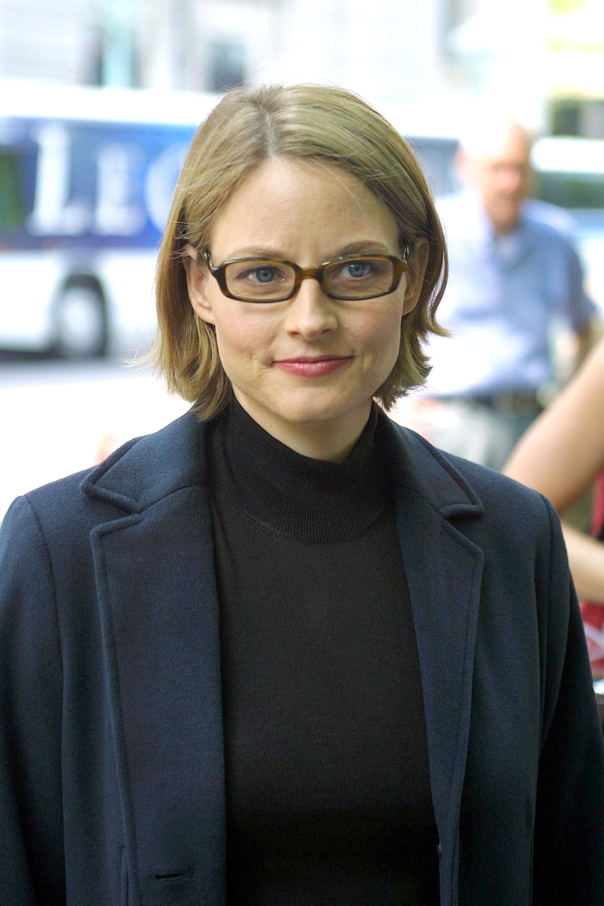 Jodie Foster wearing glasses, a black turtleneck, and a dark coat, photographed outdoors